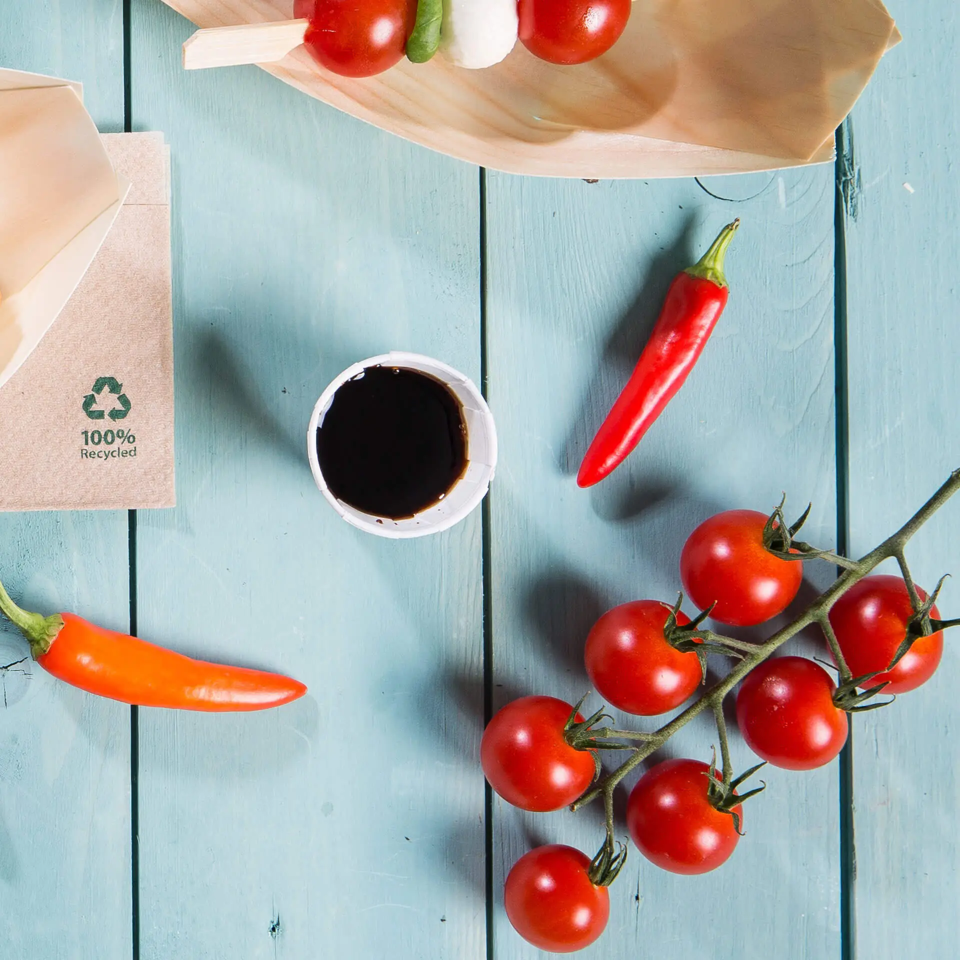 Obraz drewnianego talerza z pomidorkami cherry, papryczkami chili, małym białym kubeczkiem papierowym z ciemnym sosem i brązowym opakowaniem z symbolem recyklingu.