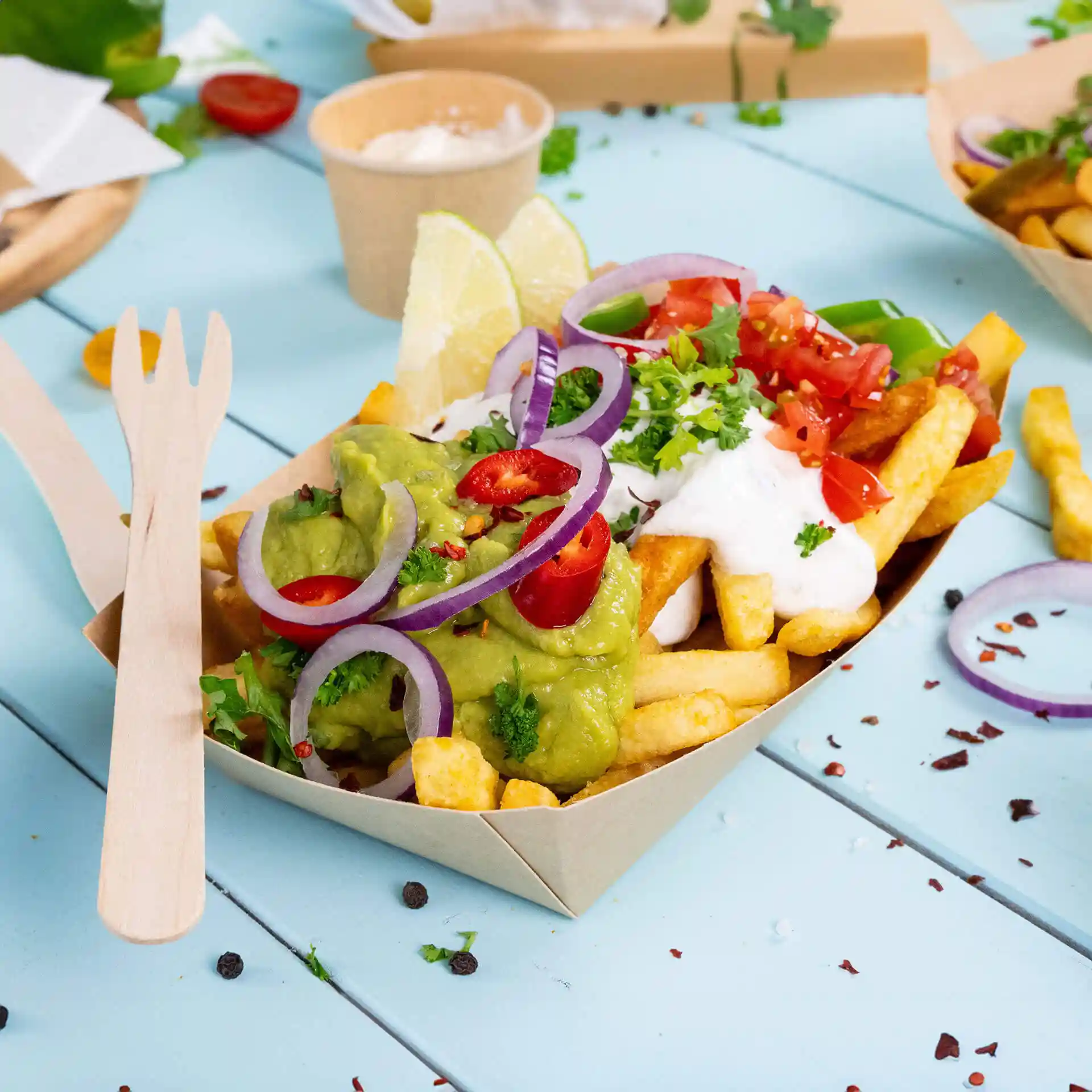 Głęboka miska z tektury wypełniona frytkami, guacamole, śmietaną, pokrojonymi pomidorami i udekorowana cebulą i limonką.