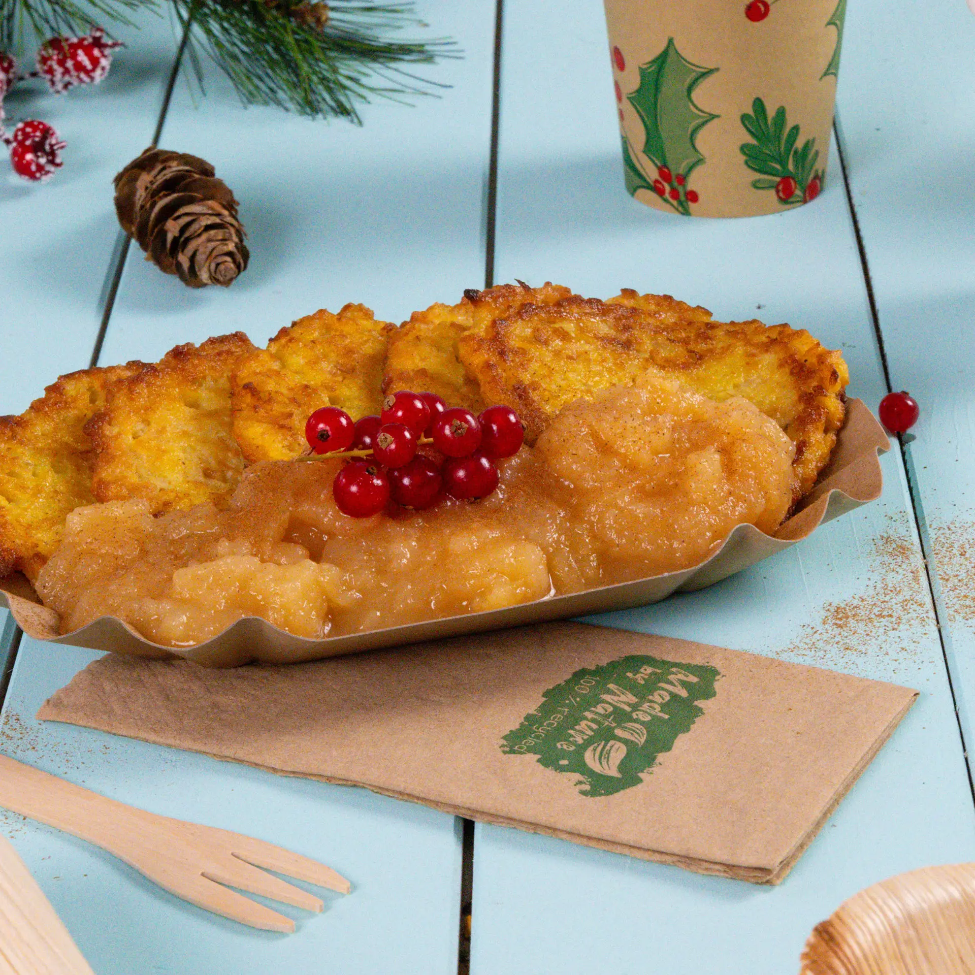 Talerz chrupiących placków ziemniaczanych podany z sosem jabłkowym, udekorowany czerwoną porzeczką, obok papierowej serwetki i elementów dekoracyjnych.