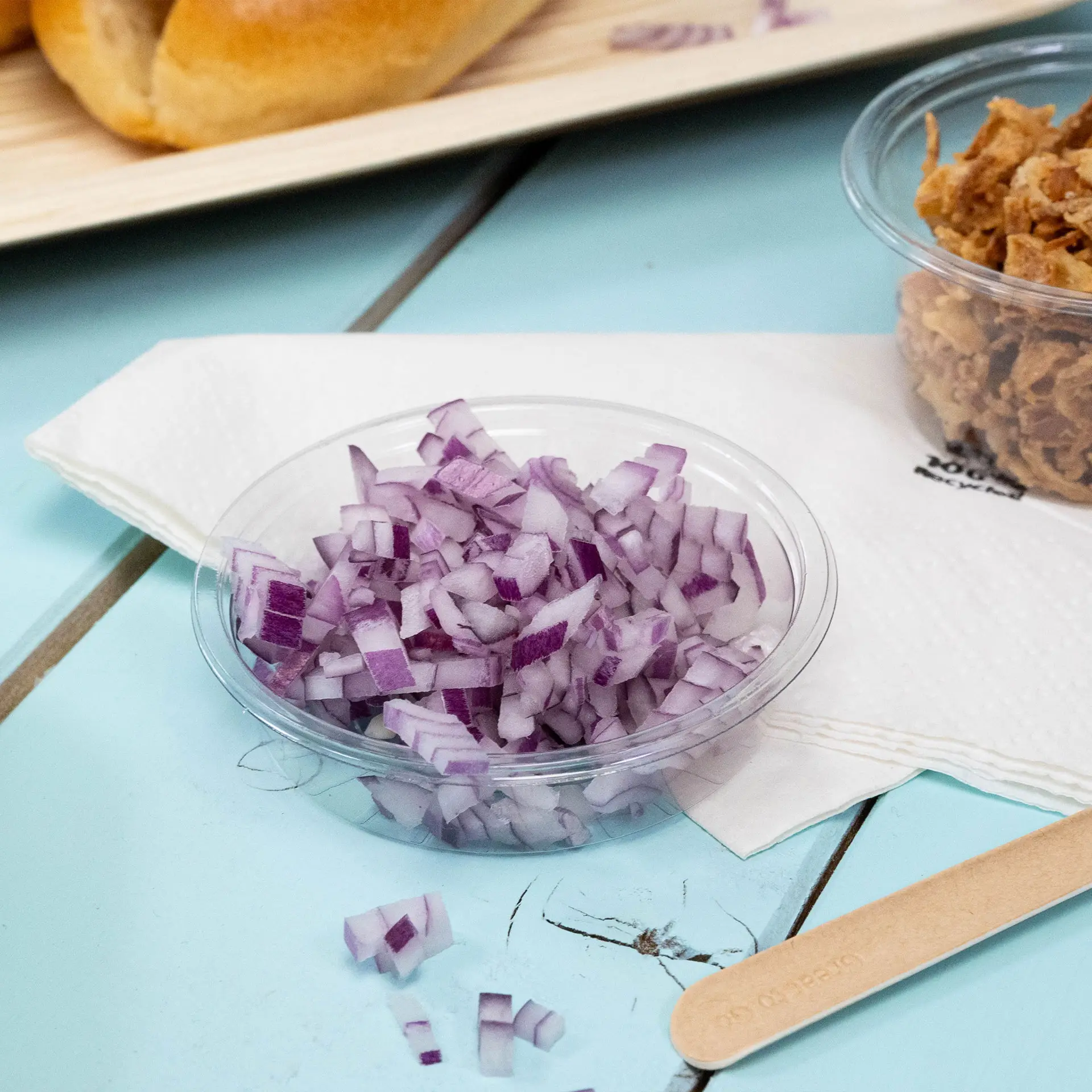 Przezroczysty plastikowy kubek z pokrojoną czerwoną cebulą, obok biała serwetka i drewniany patyczek.