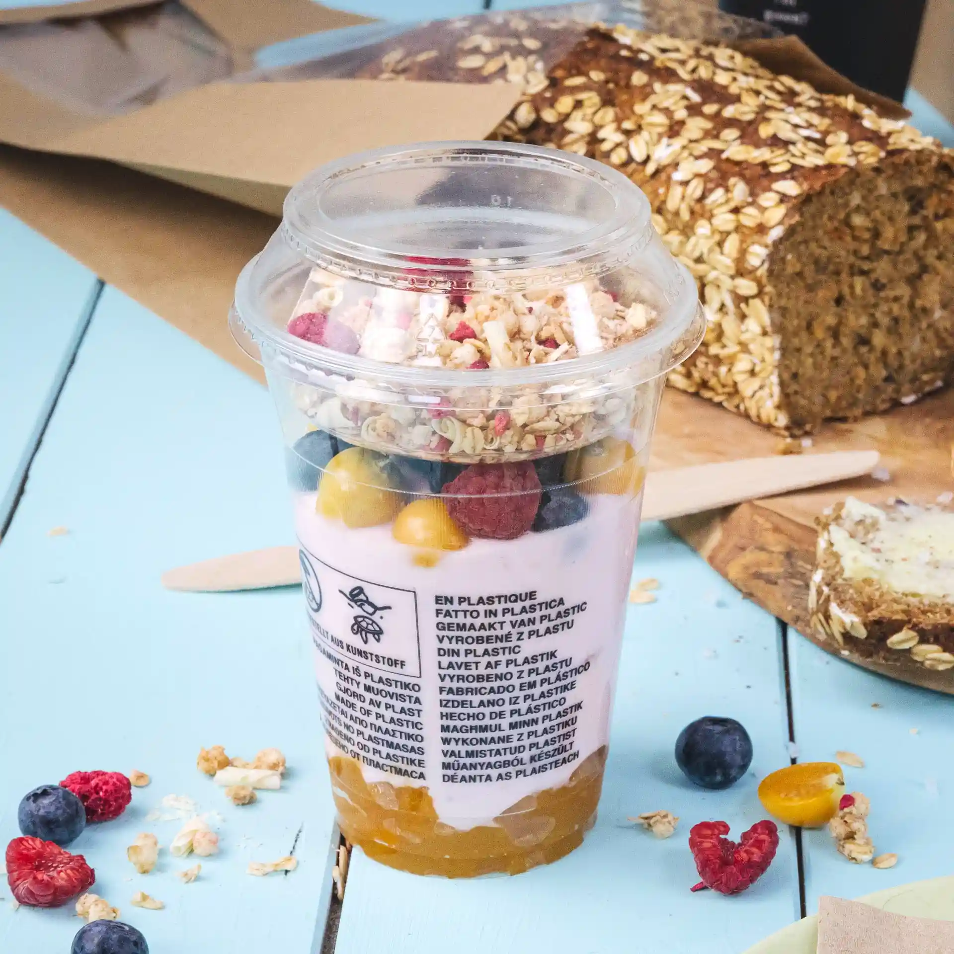 Przezroczysty kubek smoothie PLA 12 oz wypełniony jogurtem, granolą i mieszanymi owocami, na niebieskiej powierzchni.