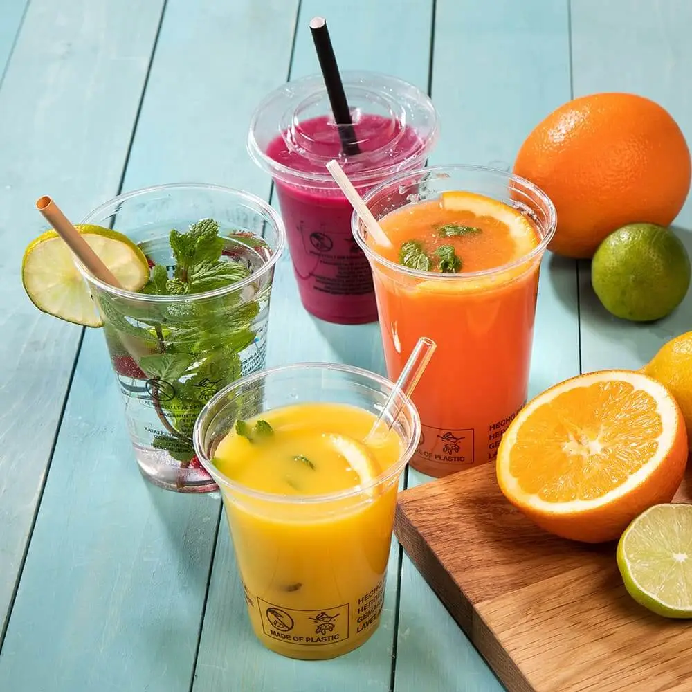 Cztery przezroczyste plastikowe kubki wypełnione kolorowymi napojami, w tym sokiem pomarańczowym i różowym smoothie, obok świeżych owoców.