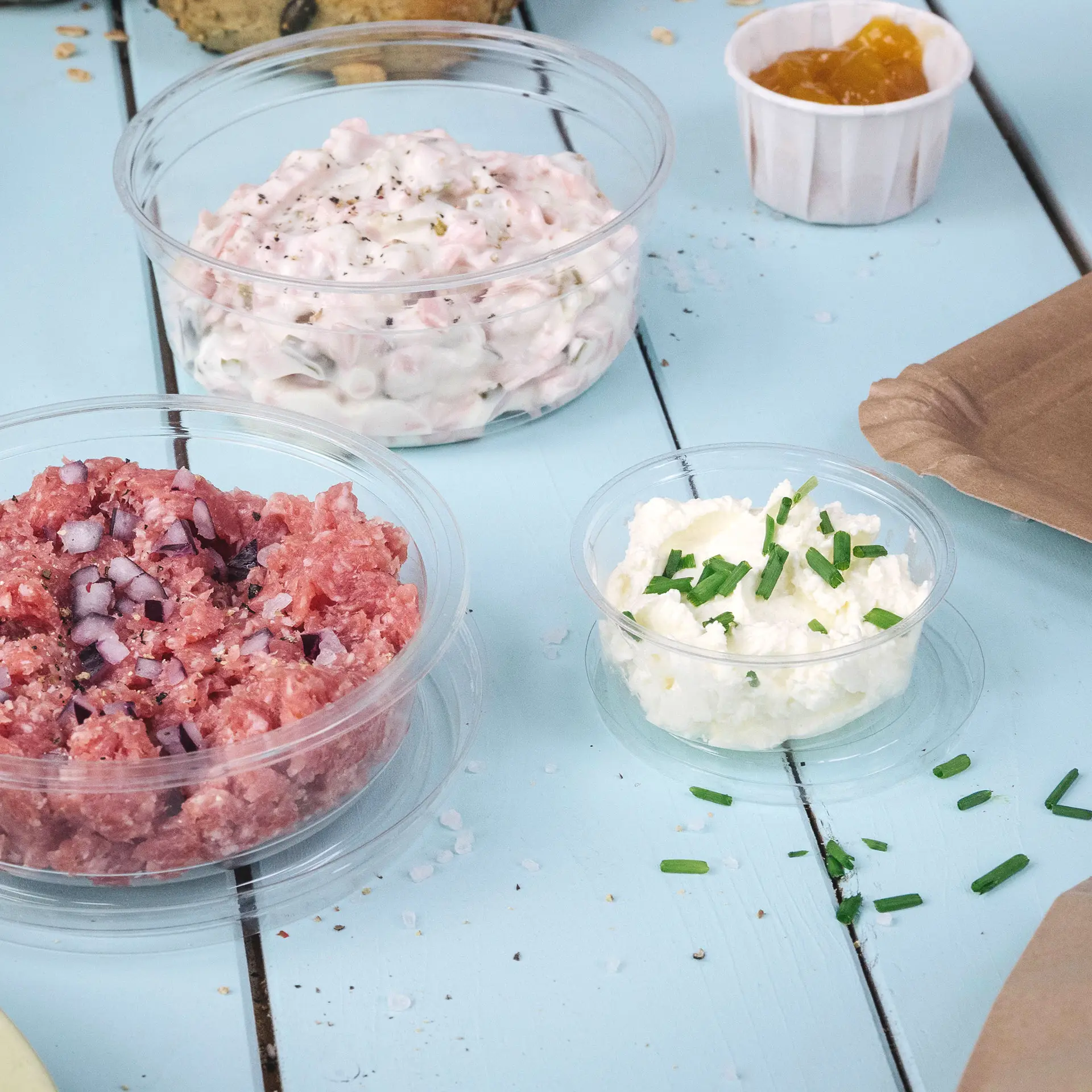 Trzy przezroczyste kubki porcyjne PLA zawierające różne produkty spożywcze, w tym różowy dip z cebulą, biały dip z szczypiorkiem i mały kubek z dżemem owocowym.