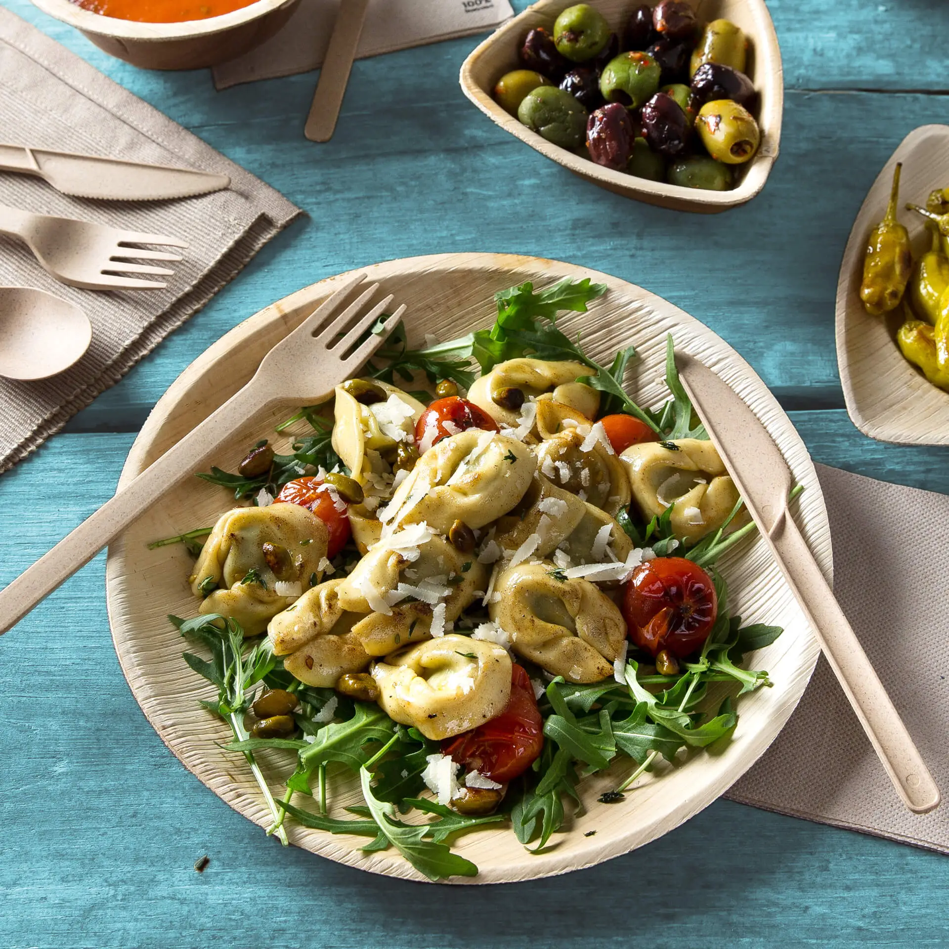 Okrągły talerz z liści palmowych wypełniony sałatką, tortellini, pomidorkami koktajlowymi i rukolą, obok drewniany widelec.