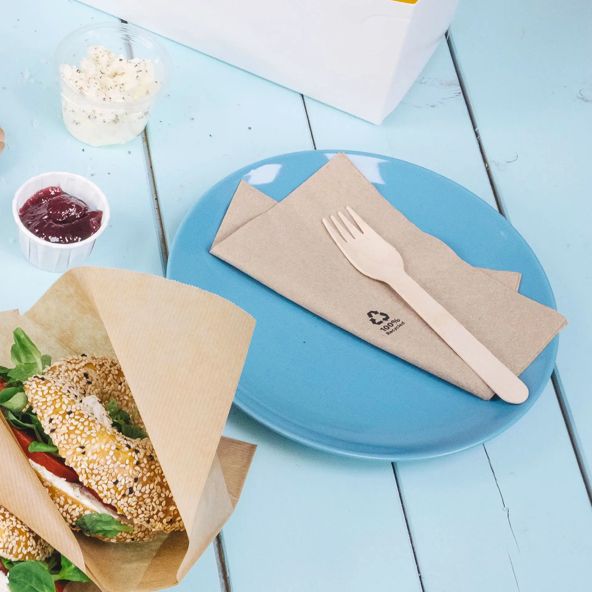 Niebieski talerz z złożonymi brązowymi serwetkami i drewnianym widelcem; sezamowa bagel z dodatkami obok, dżem w kubku.