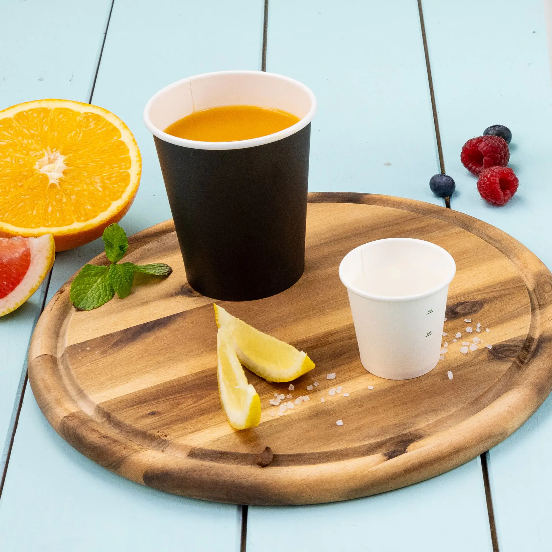 Czarny papierowy kubek pokryty PLA, wypełniony pomarańczowym płynem, w towarzystwie małego białego kubka, plasterków cytryny, mięty i owoców na drewnianej tacy.