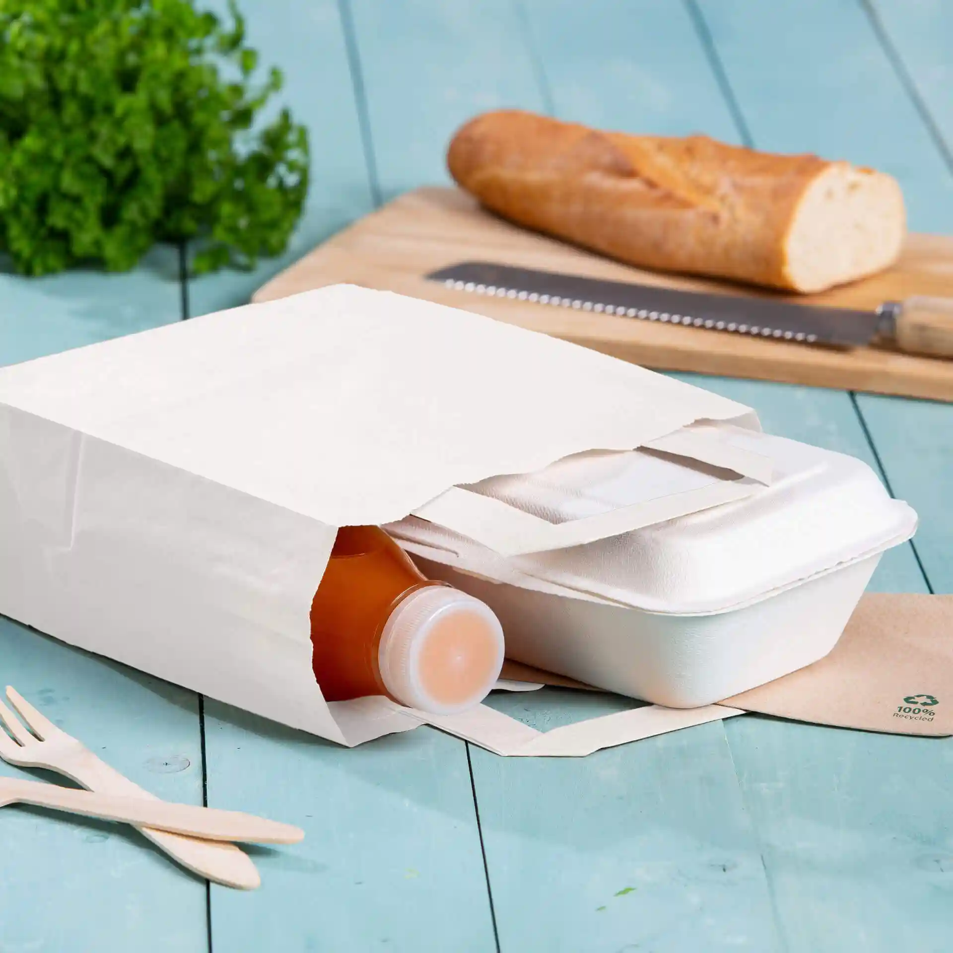 Biała torba papierowa kraft z butelką i pojemnikiem na wynos w środku, umieszczona na niebieskiej drewnianej powierzchni.