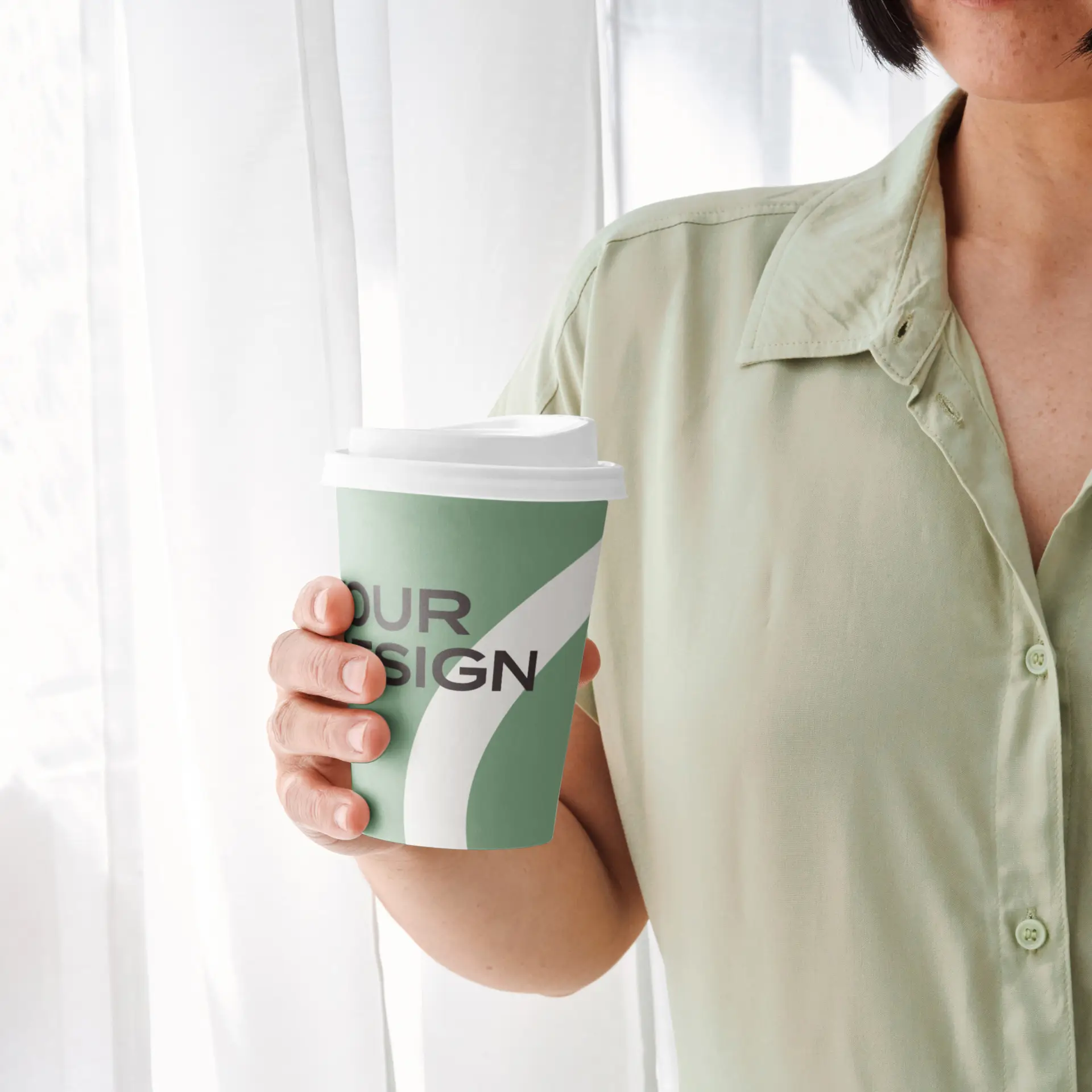 Ręczny zielony kubek papierowy z białą pokrywką, z logo i grafiką; osoba w tle ma na sobie jasnoniebieską koszulę.