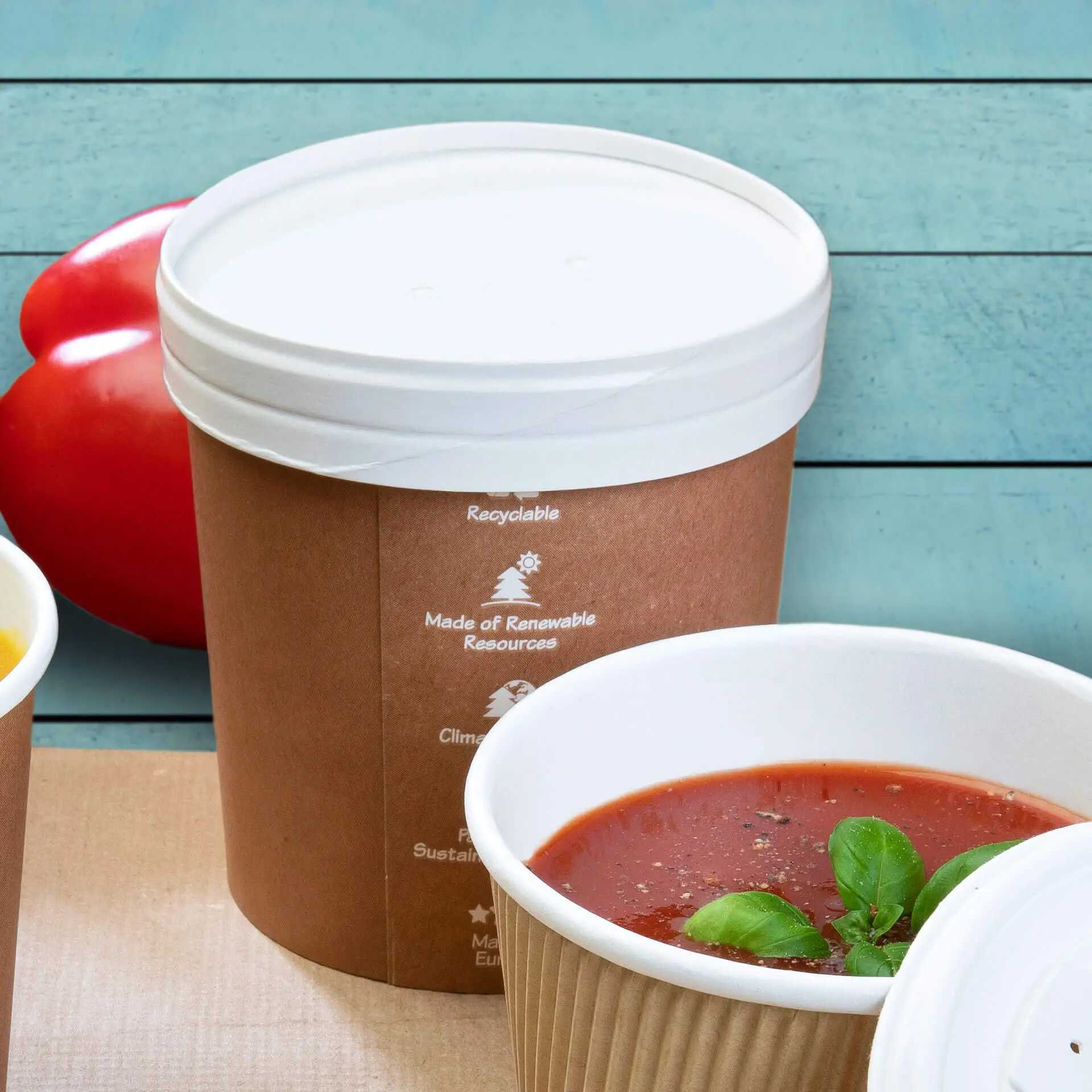 Kartonowy kubek na zupę z białą pokrywką membranową, pokryty bio-PE; dodatek bazylii w pobliskim kubku; czerwona pomidor na tle.