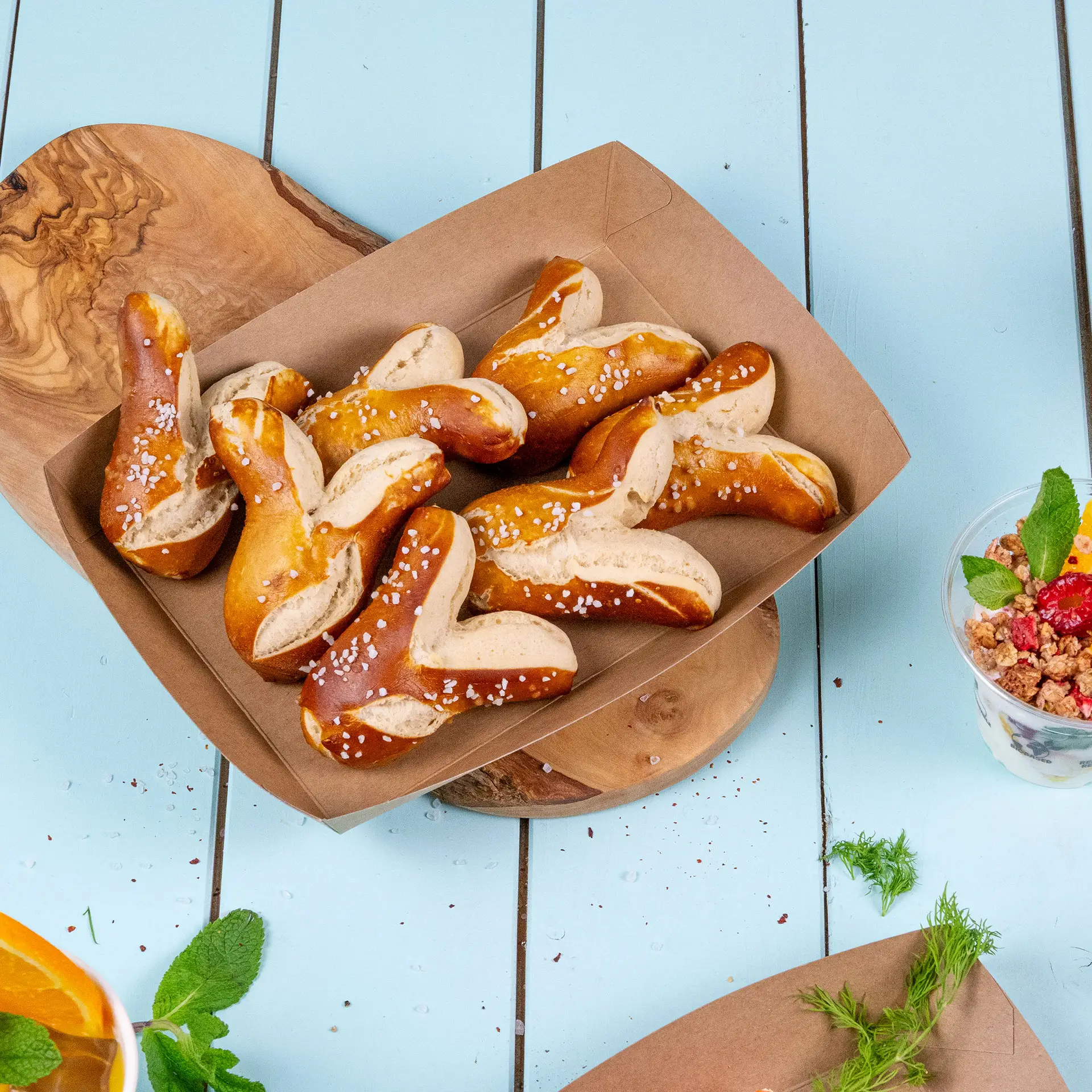 Brąska tektura spożywcza wypełniona precle, posypana grubą solą, umieszczona na drewnianej desce.