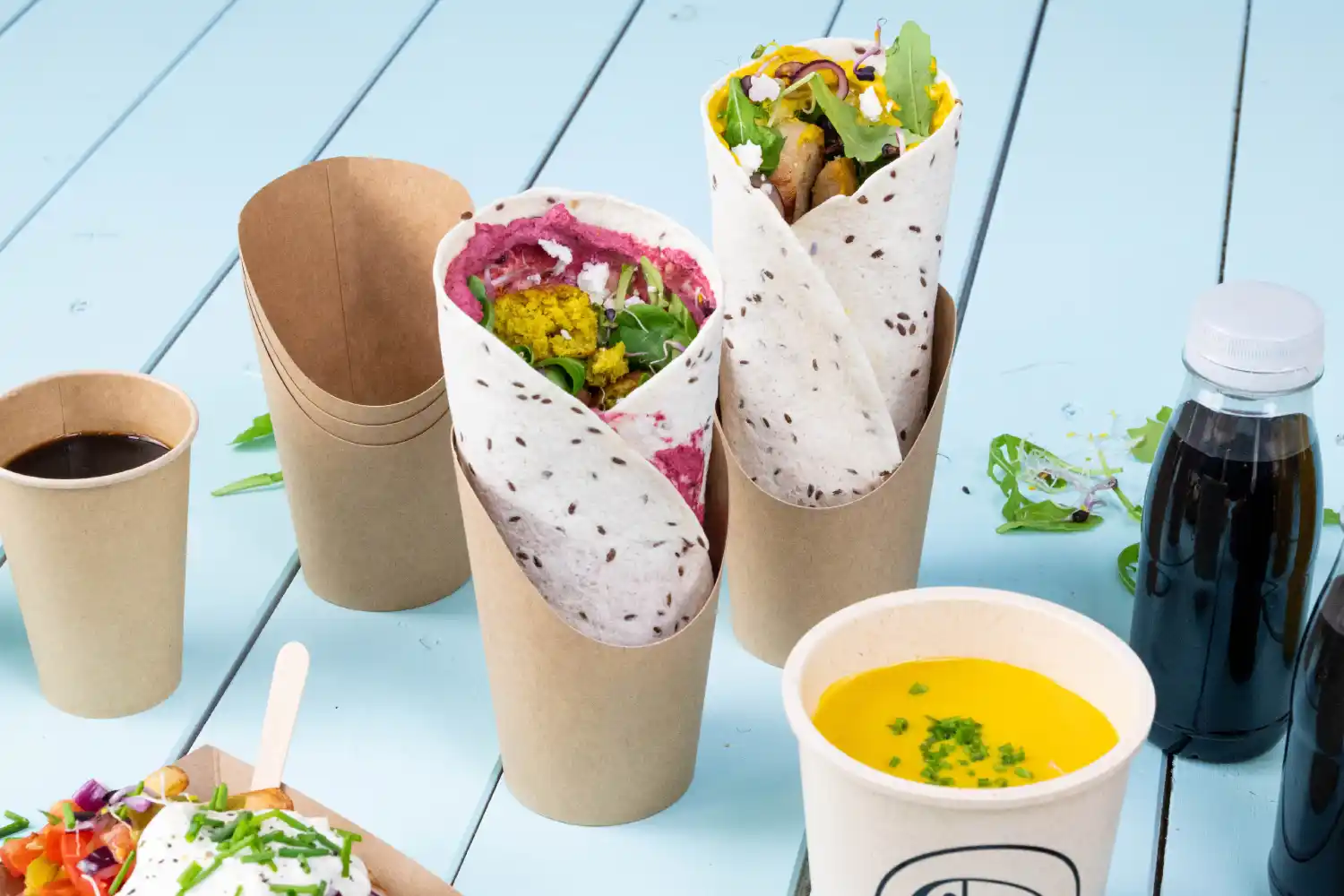 Dwa zapakowane produkty w kubkach, jeden z różowym nadzieniem i zieleniną, drugi żółty, obok filiżanki kawy i zupy.