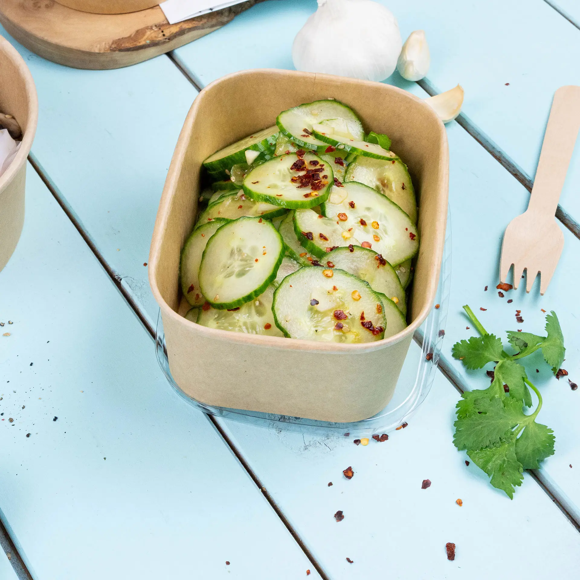 Brąski pojemnik na żywność wypełniony pokrojonymi ogórkami, posypany płatkami chili, na jasnoniebieskiej drewnianej powierzchni.