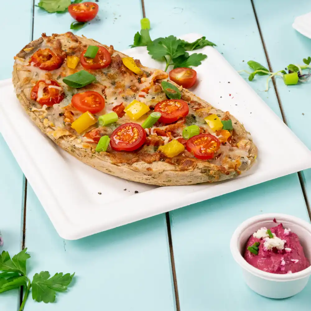 Pieczeń płaskiego chleba z pomidorkami koktajlowymi, zieloną cebulą i papryką, podana na białym talerzu z dodatkiem dipu.