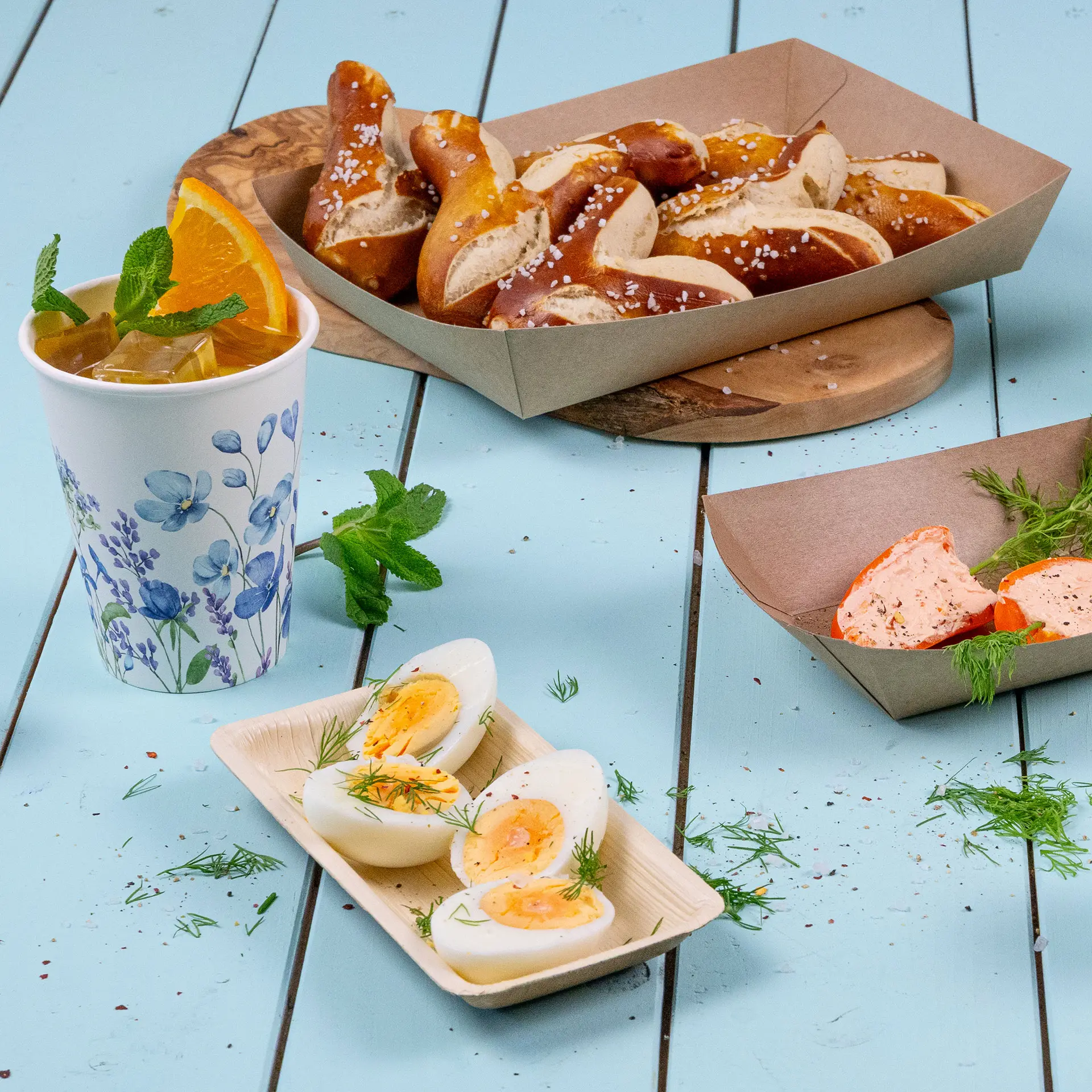 Prostokątna miska z liści palmy z gotowanymi jajkami, przyozdobiona koperkiem; kubek mrożonej herbaty z miętą i pomarańczą; precle w pudełku obok.