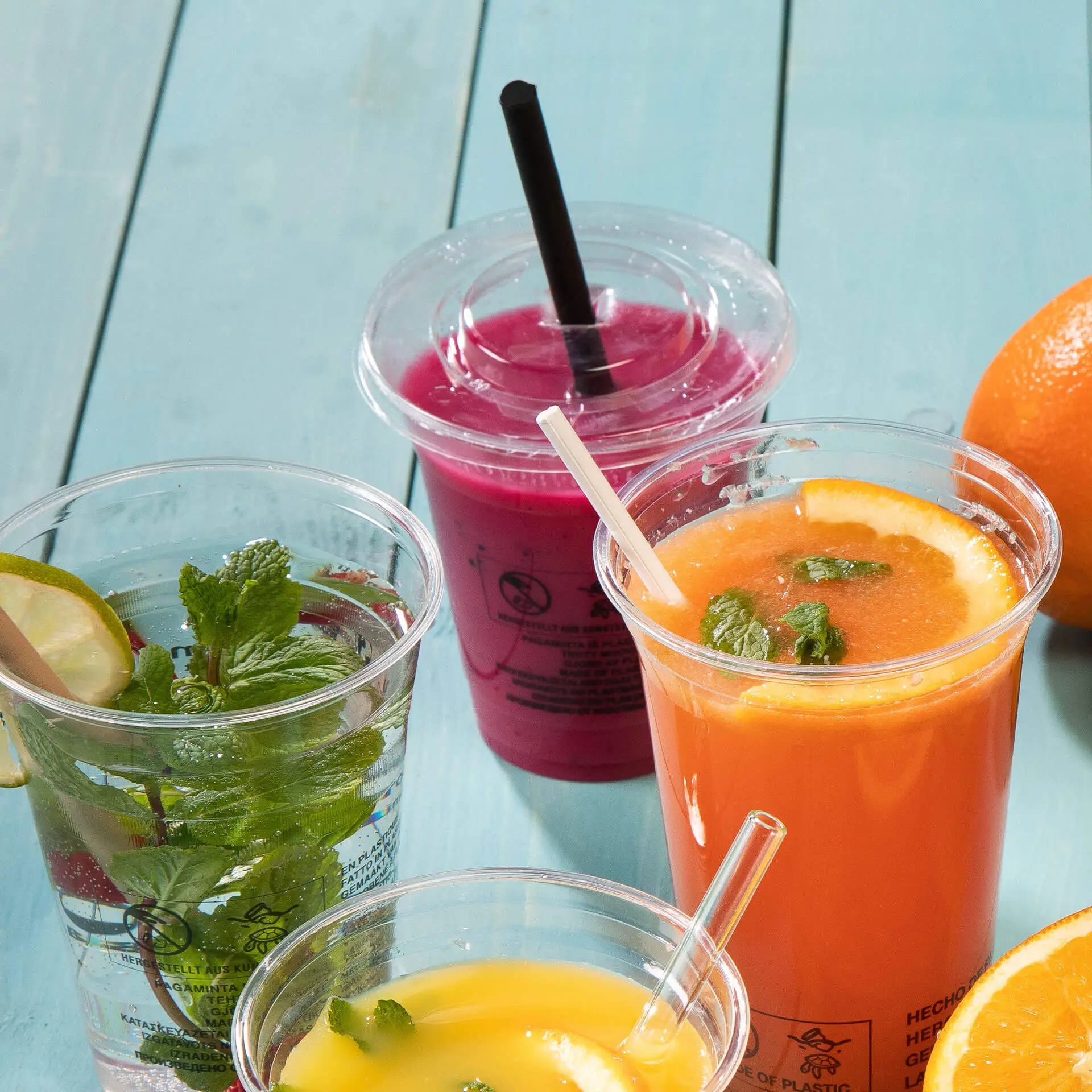 Trzy przezroczyste kubki z napojami: jeden z miętą i limonką, napój pomarańczowy z miętą i różowy napój ze słomką; w tle pomarańcza.