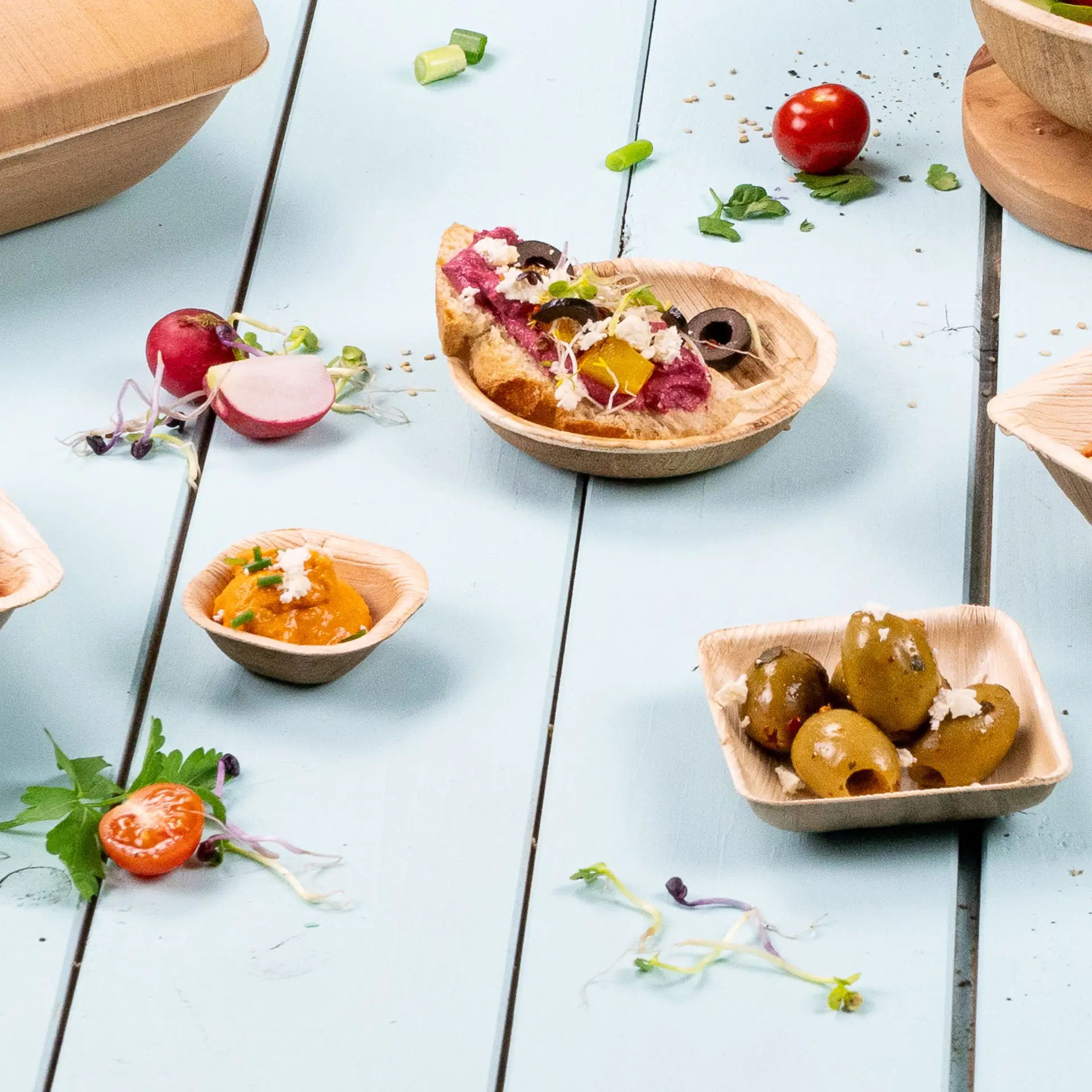 Mini miski z liści palmy, 80 ml, ułożone na niebieskim stole z różnymi produktami, takimi jak oliwki, dipsy i warzywa.