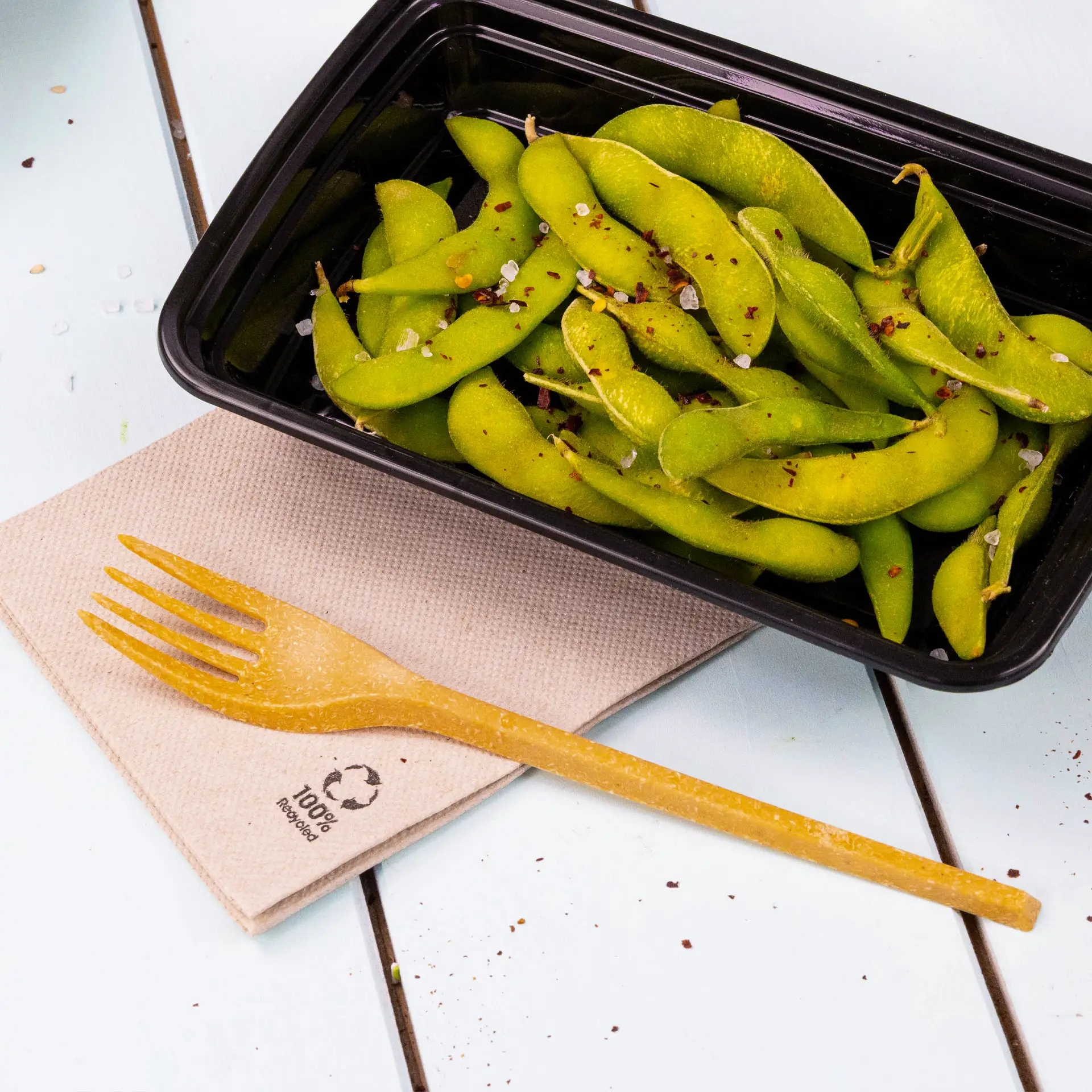 Obraz świeżych nasion edamame w czarnym pojemniku, w towarzystwie biodegradowalnego widelca na recyklingowym serwetce papierowej.