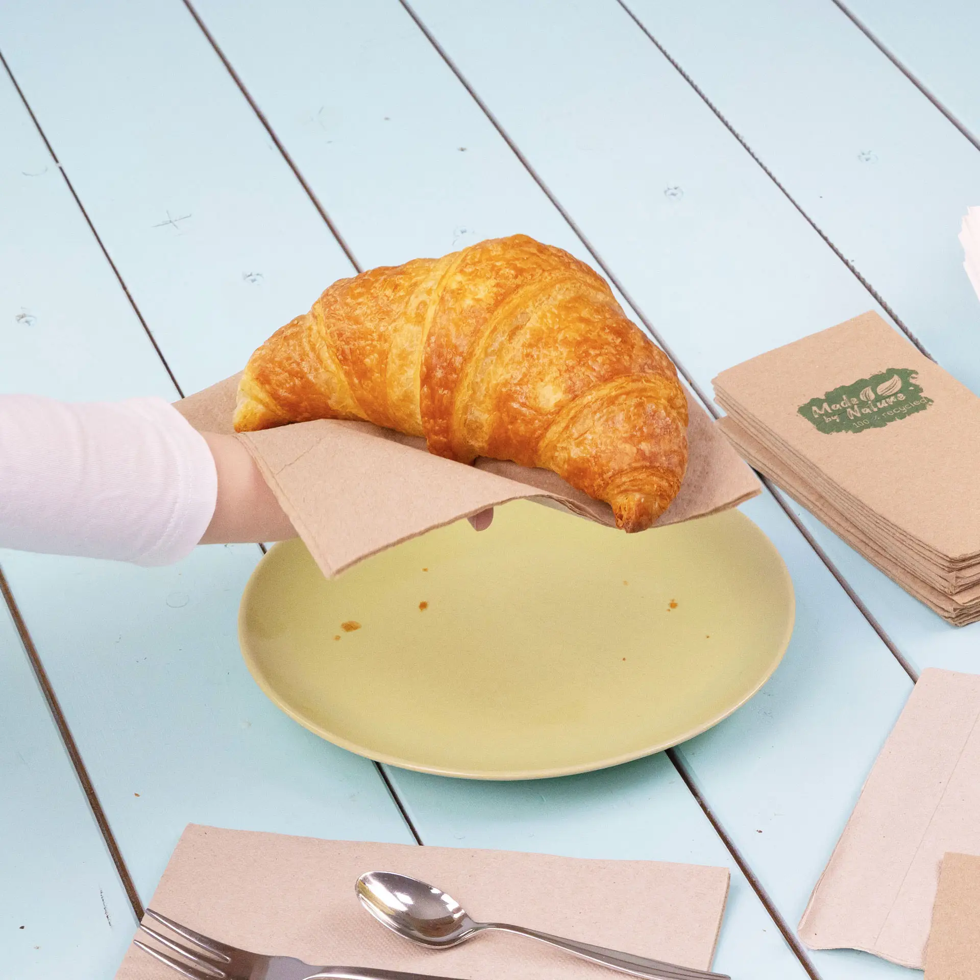 Ręka trzyma croissanta na papierowym serwetce nad żółtym talerzu, z dodatkowymi serwetkami i sztućcami w pobliżu.