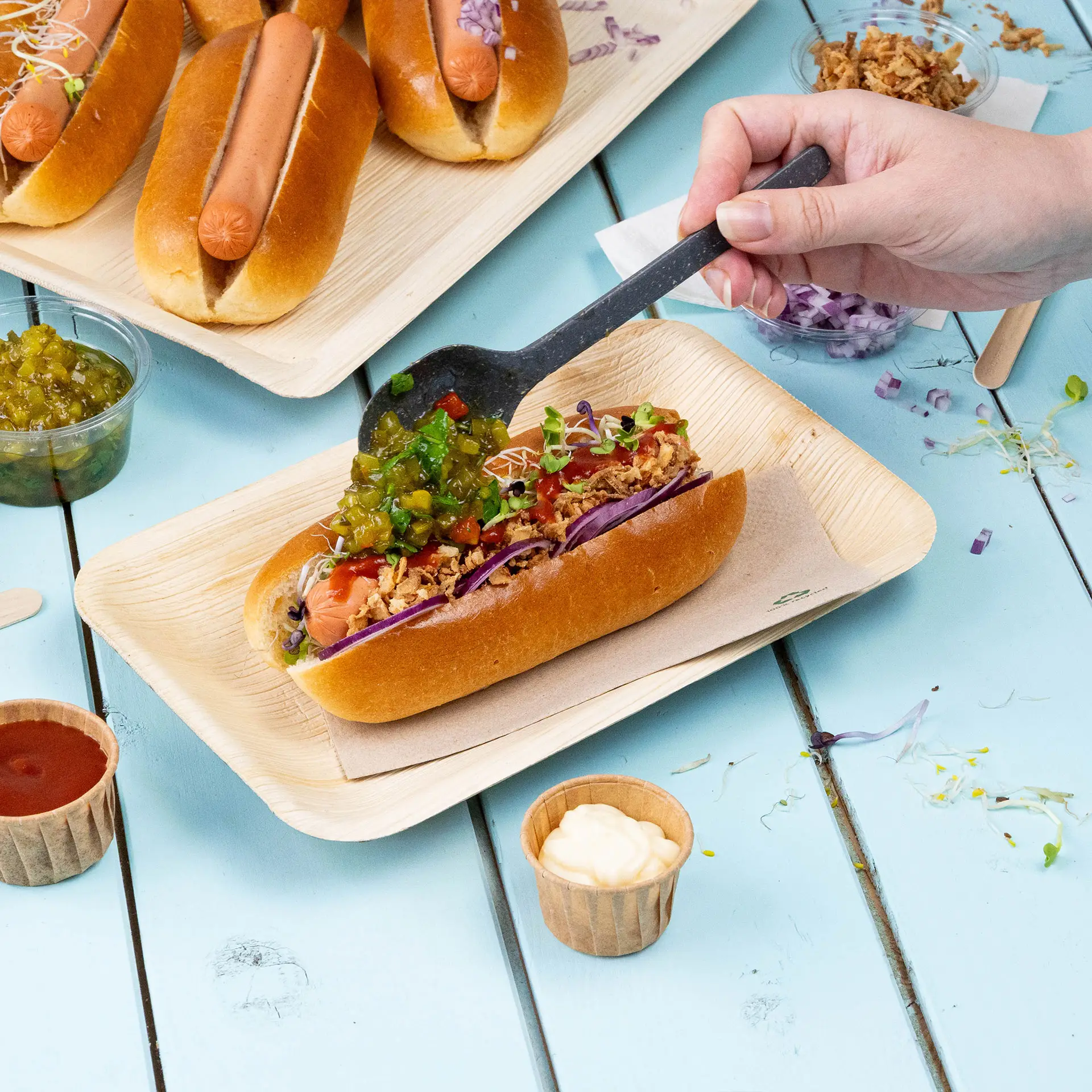 Ręka trzyma łyżkę, dodając dodatki do hot doga na talerzu z liści palmowych, widoczne są dodatkowe hot dogi i sosy.