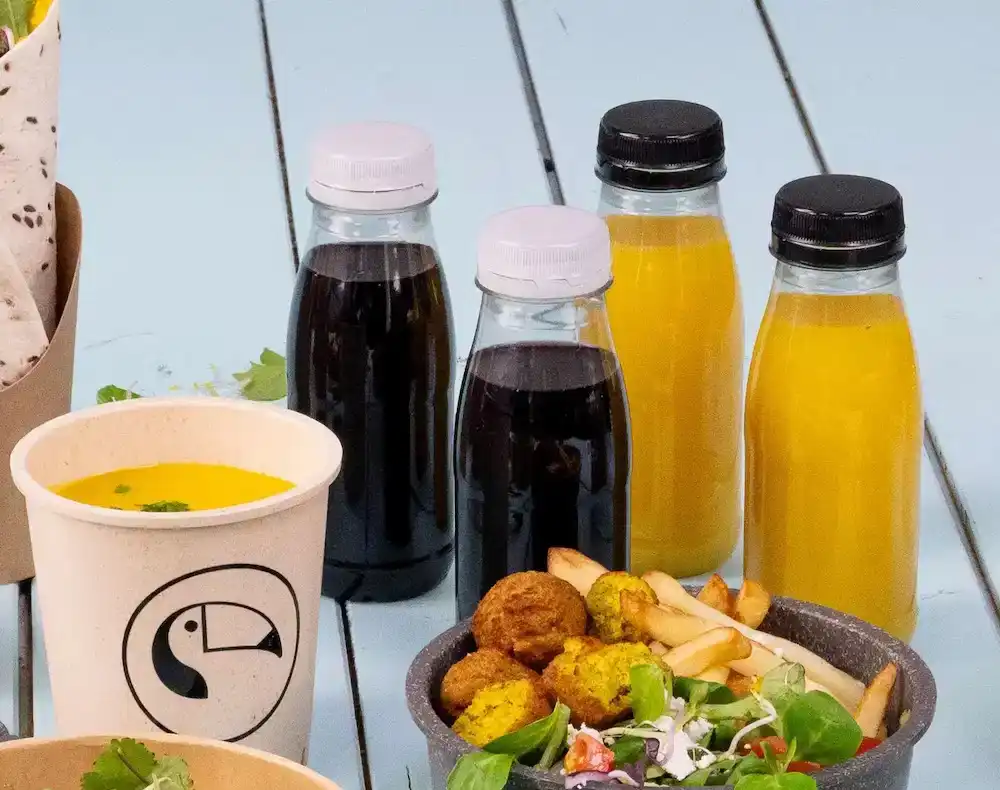 Różnorodne produkty spożywcze, w tym butelki z plastiku rPET z różnymi napojami, kubek zupy i miska sałatki, frytek i falafela.