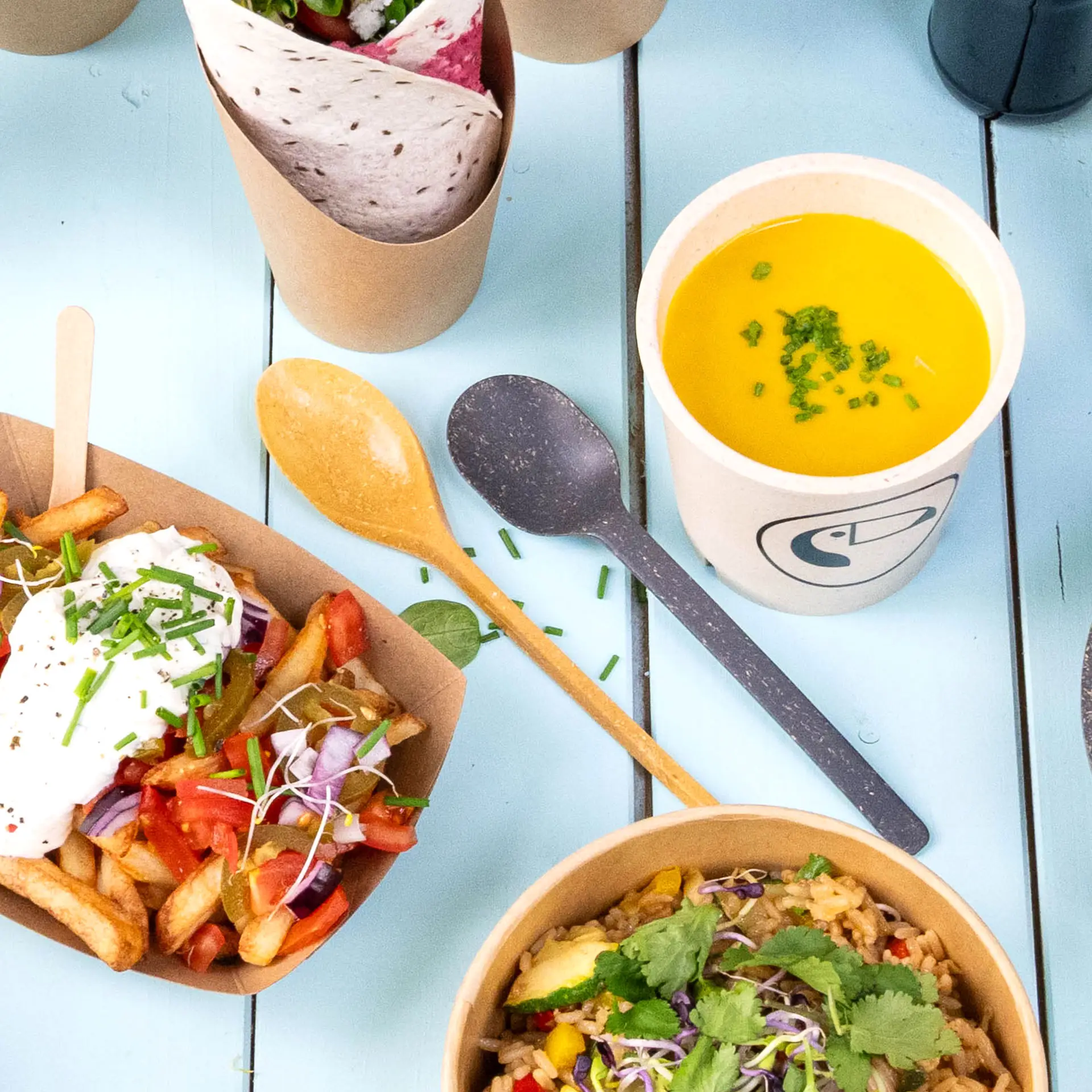 Jedna wielorazowa łyżka w kolorze karmelowym z biokompozytu; zawiera różne artykuły spożywcze i żółty kubek do zupy z szczypiorkiem.