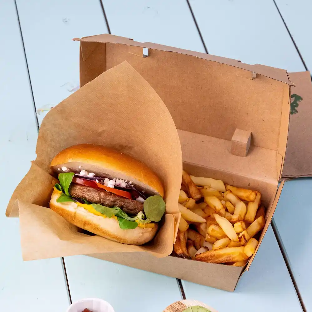 Burger w papierowym opakowaniu z świeżymi warzywami obok pudełka frytek na niebieskiej powierzchni.