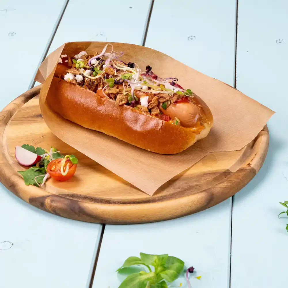 Hot dog w miękkiej bułce, pokryty chrupiącą cebulą, kiełkami i udekorowany pomidorkami koktajlowymi oraz rzodkiewką na drewnianym talerzu.