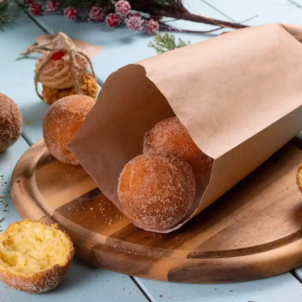 Brązka papierowa torba zawiera kilka okrągłych donuts posypanych cukrem pudrem na drewnianej desce do krojenia, jeden donut częściowo pokrojony.