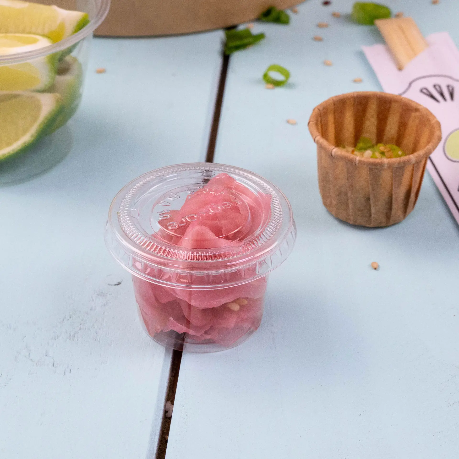 Przezroczysty plastikowy kubek z różowym marynowanym imbirem na jasnoniebieskiej powierzchni, w tle brązowy papierowy kubek i plasterki limonki.