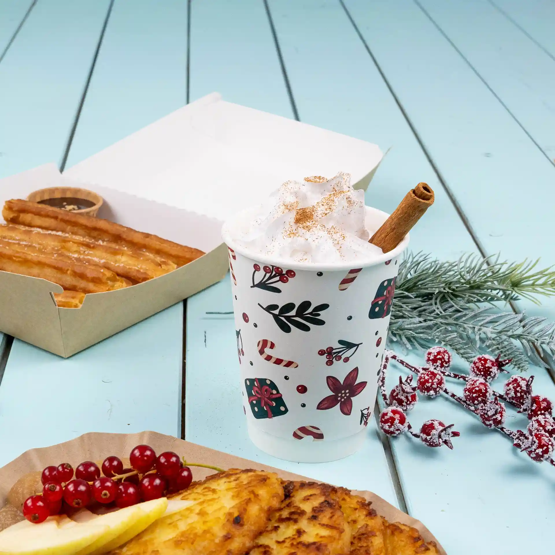 Podwójna szklanka z zimowym wzorem, wypełniona bitą śmietaną i laską cynamonu, obok churros i owoców.
