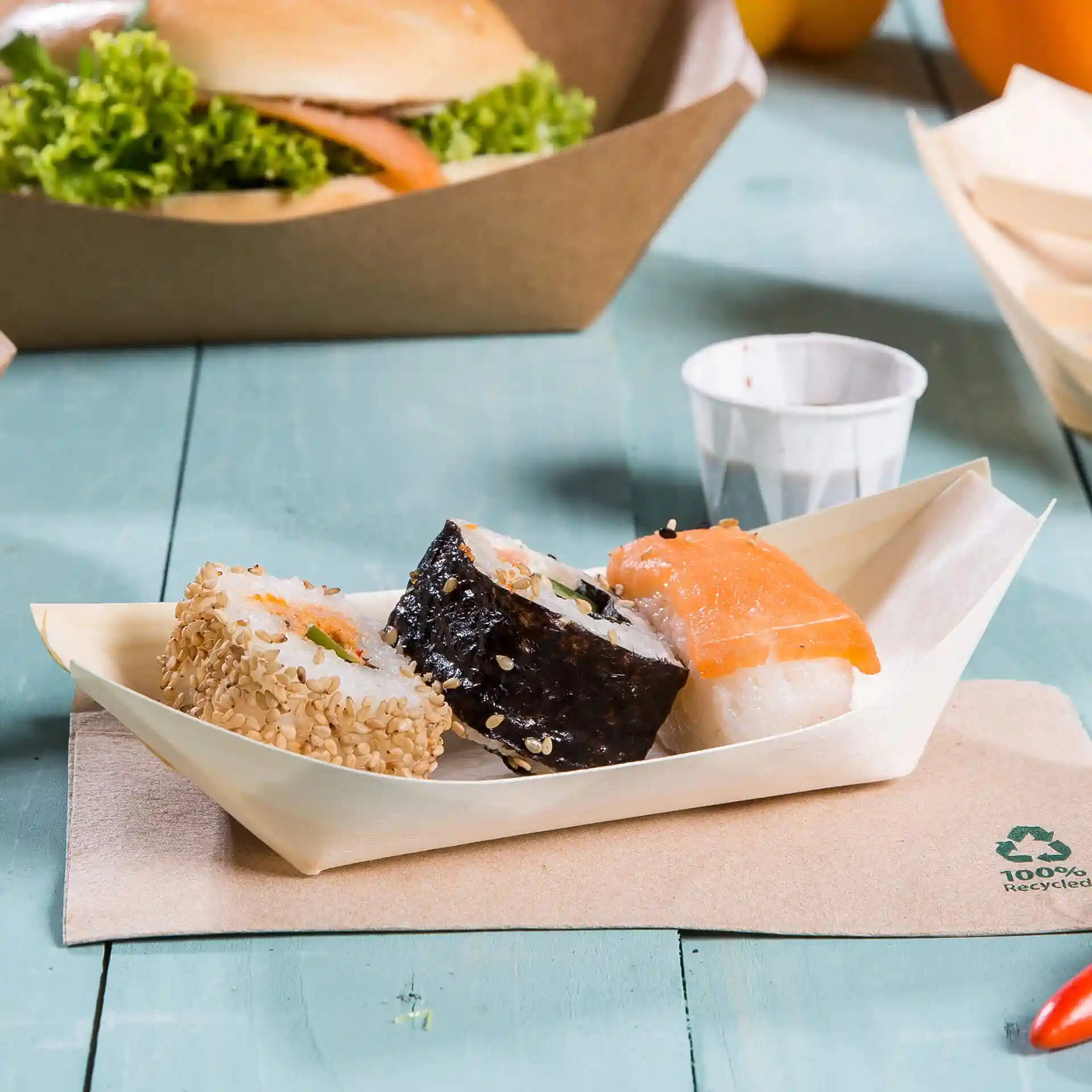 Trzy kawałki sushi podane na drewnianej tacy w kształcie łodzi na jasnoniebieskiej powierzchni.