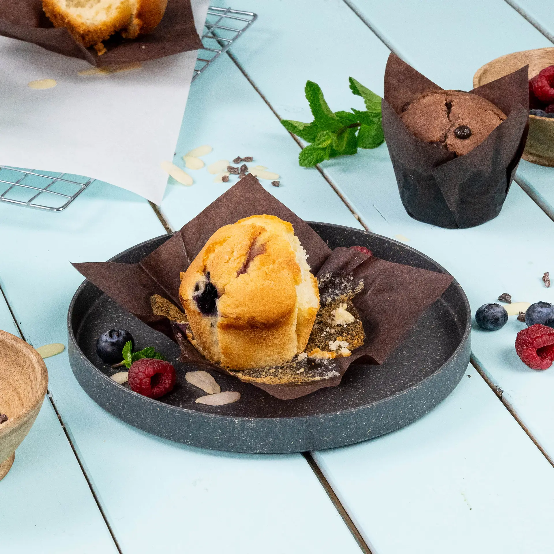 Muffin w brąskim papierowym kubku odpornym na tłuszcz, z borówkami i malinami na szarym talerzu, na jasnoniebieskim tle.