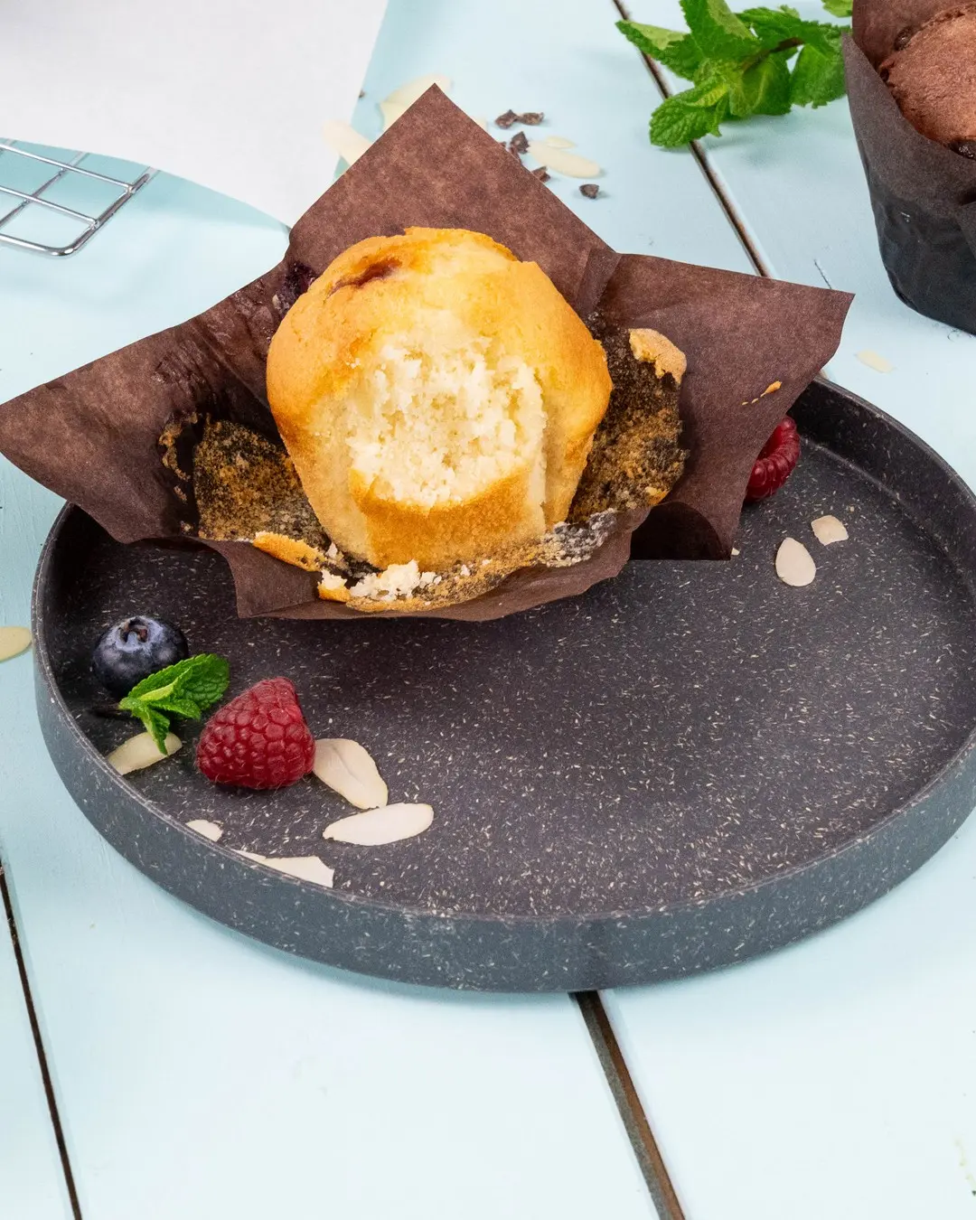 Muffin w brązowym papierowym opakowaniu na ciemnym talerzu, otoczony malinami, borówkami, liśćmi mięty i plasterkami migdałów.
