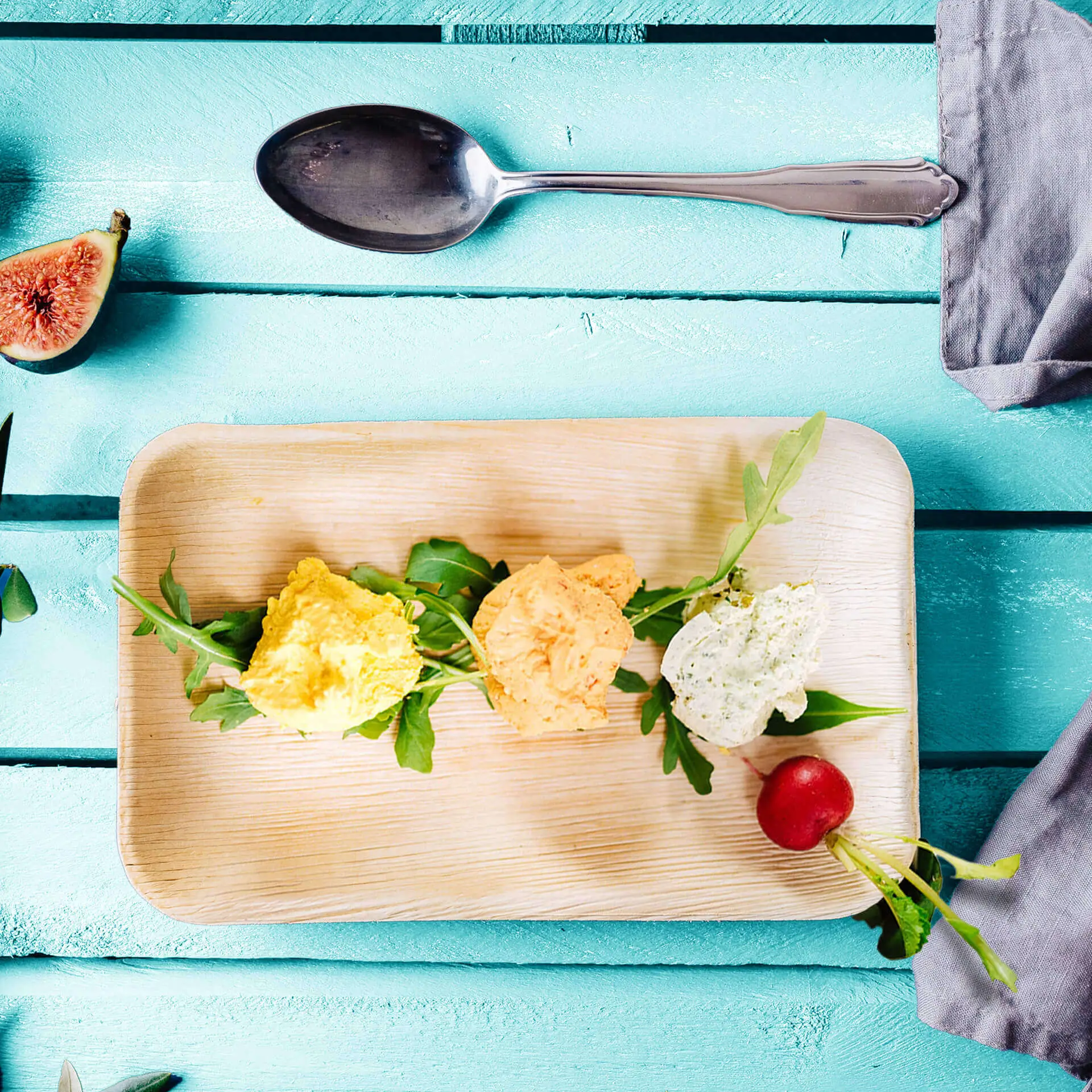 Trzy kolorowe kulki hummusu na rukoli, podane na talerzu z liścia palmy na niebieskim drewnianym tle, z łyżką w pobliżu.