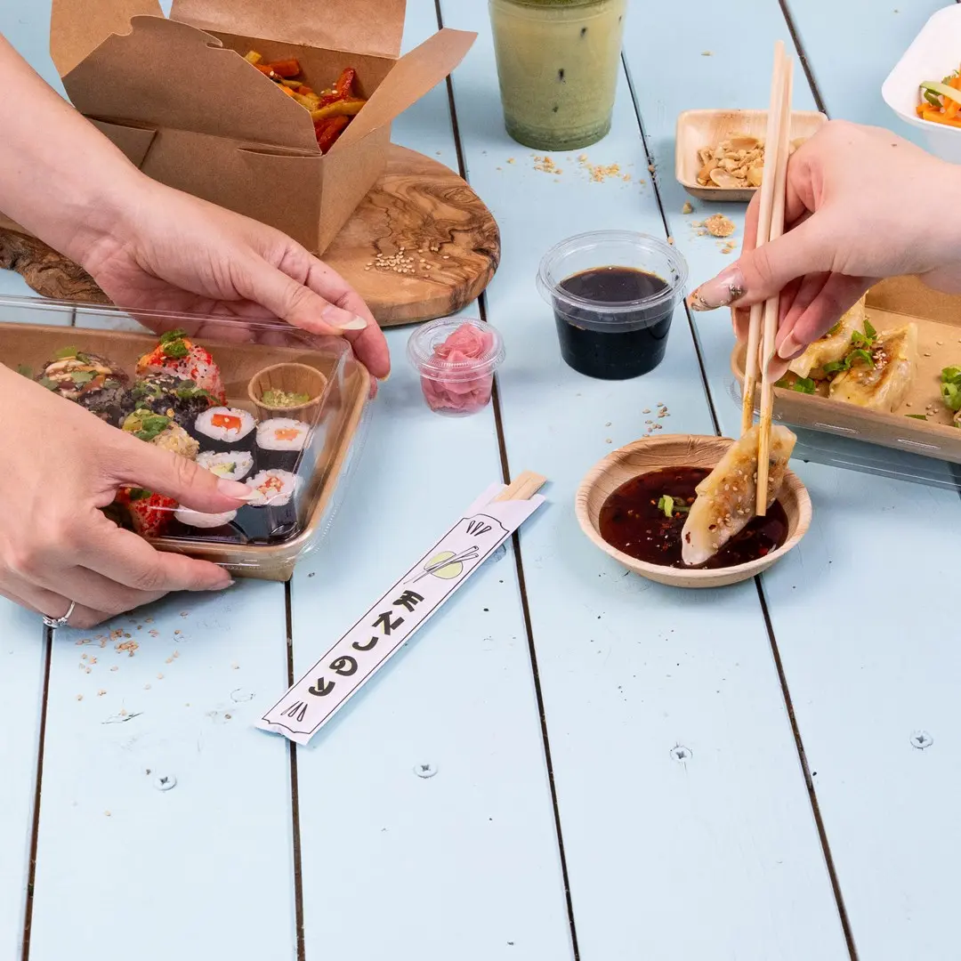 Ręce trzymają sushi w przezroczystym pojemniku, z sosem, marynowanym imbirem i pałeczkami na niebieskim stole.