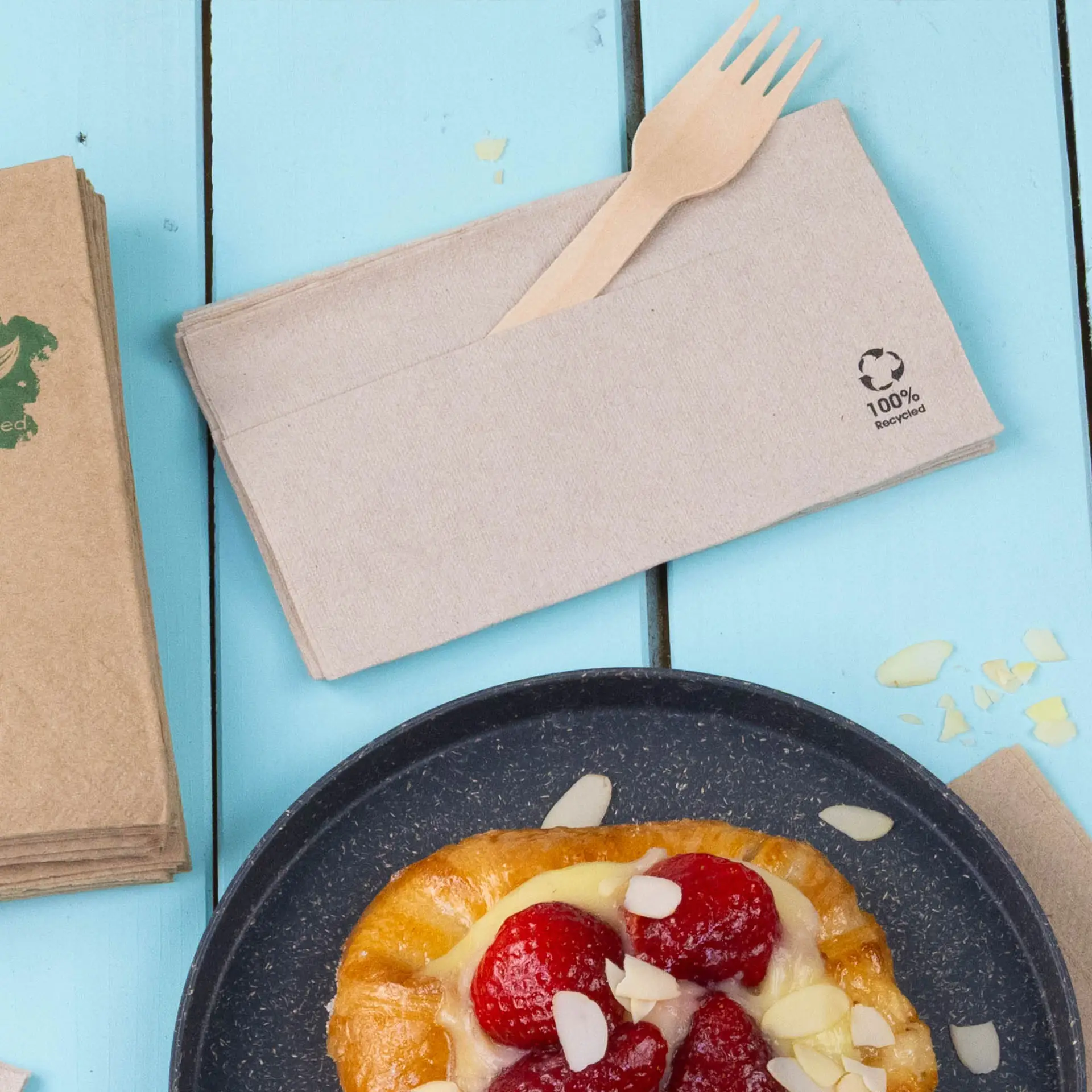 Serwetka z recyklingowego papieru z drewnianym widelcem, umieszczona obok ciastka na niebieskiej powierzchni.