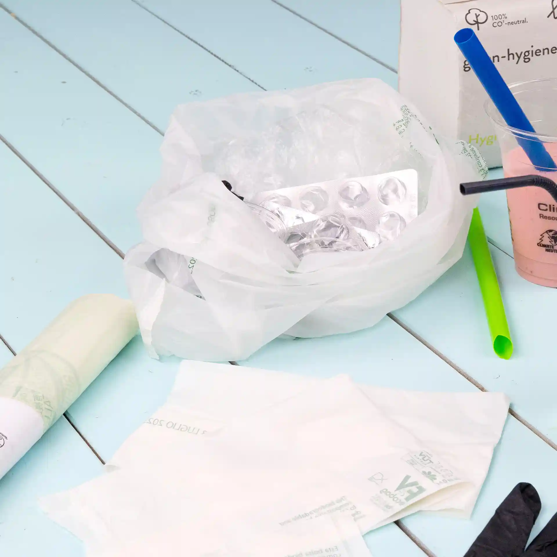 Przezrożny worek bio z zawartością, w tym opakowanie blistrowe, plastikowe słomki i jednorazowe przedmioty na niebieskiej powierzchni.