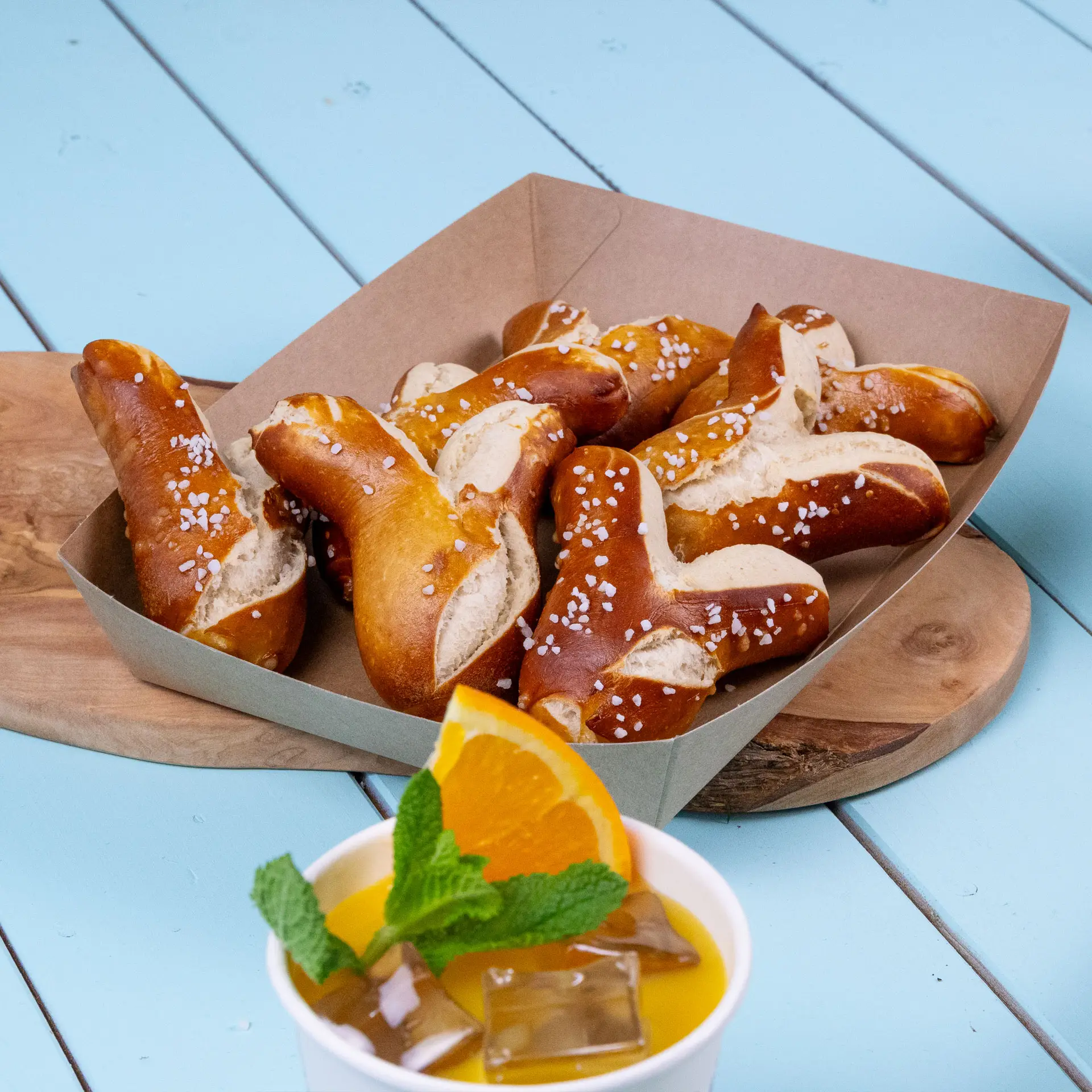 BrąTray kartonowy wypełniony preclami posypanymi grubą solą, obok kubka z napojem pomarańczowym przyozdobionym liśćmi mięty.