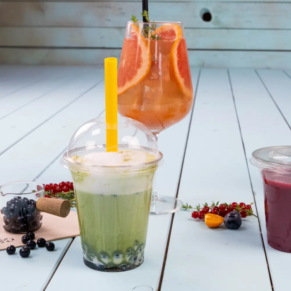 Zielony napój z żółtą słomką w przezroczystym kubku, z niebieskimi kulkami tapioki na dnie; kolorowy koktajl z plastrami grejpfruta w szklance za nim.