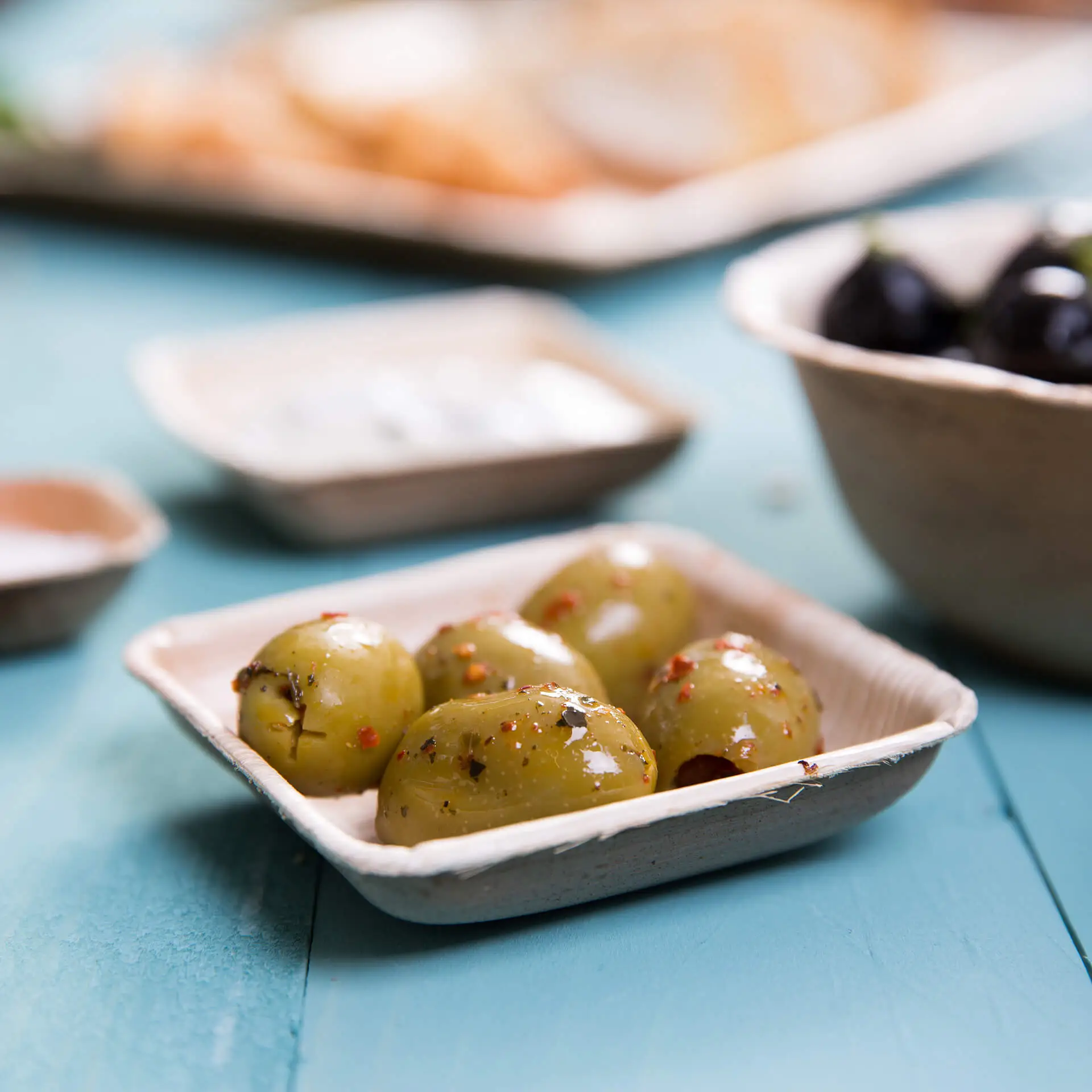 Kwadratowa miska z liści palmowych z kilkoma zielonymi oliwkami i przyprawami; lekki, ekologiczny artykuł stołowy.