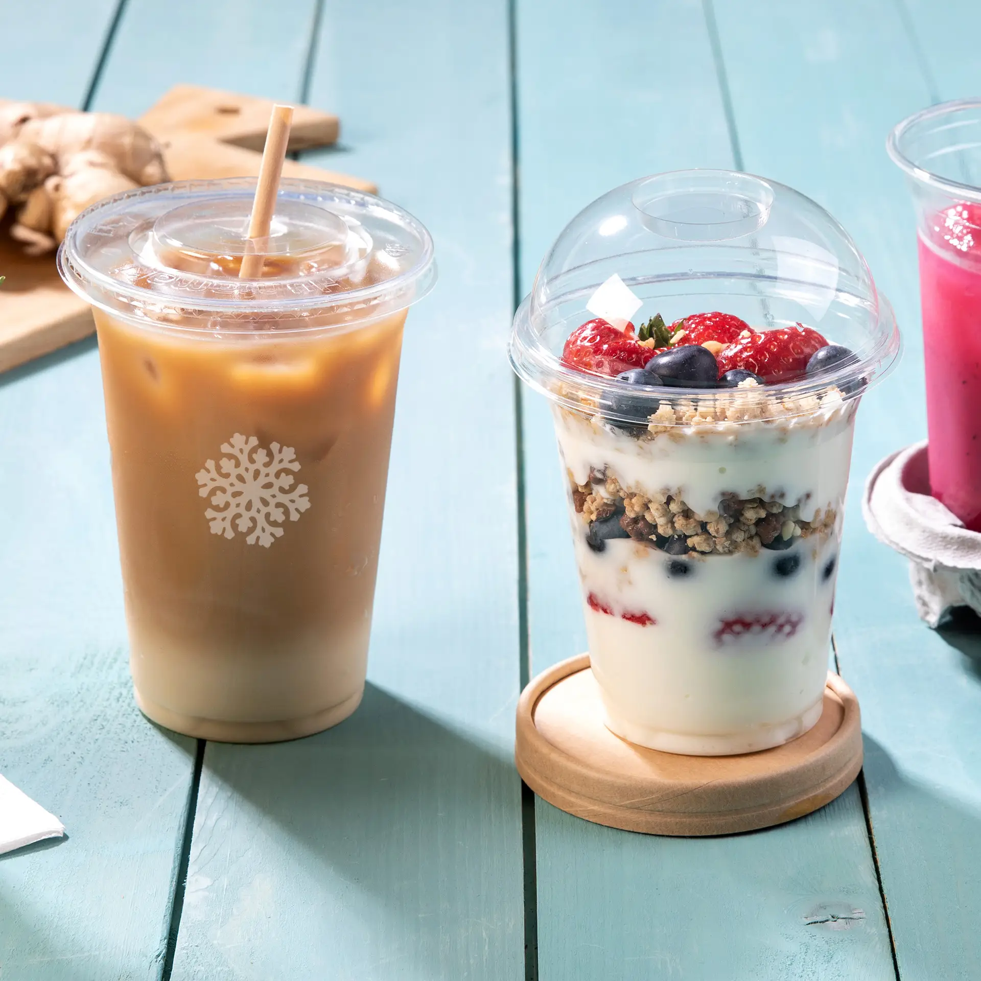 Trzy przezroczyste kubki PLA na zimne napoje: kubek z iced coffee, parfait z owocami i różowy napój, na drewnianym stole.
