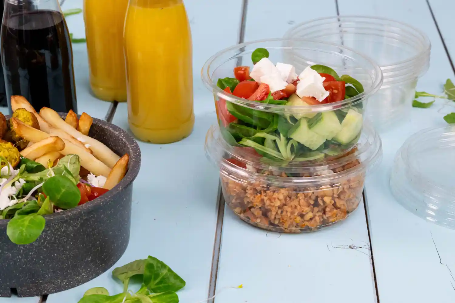 Pojemniki z sałatką i zbożami obok miski z frytkami; dwie butelki napoju w tle; jasnoniebieska drewniana powierzchnia.