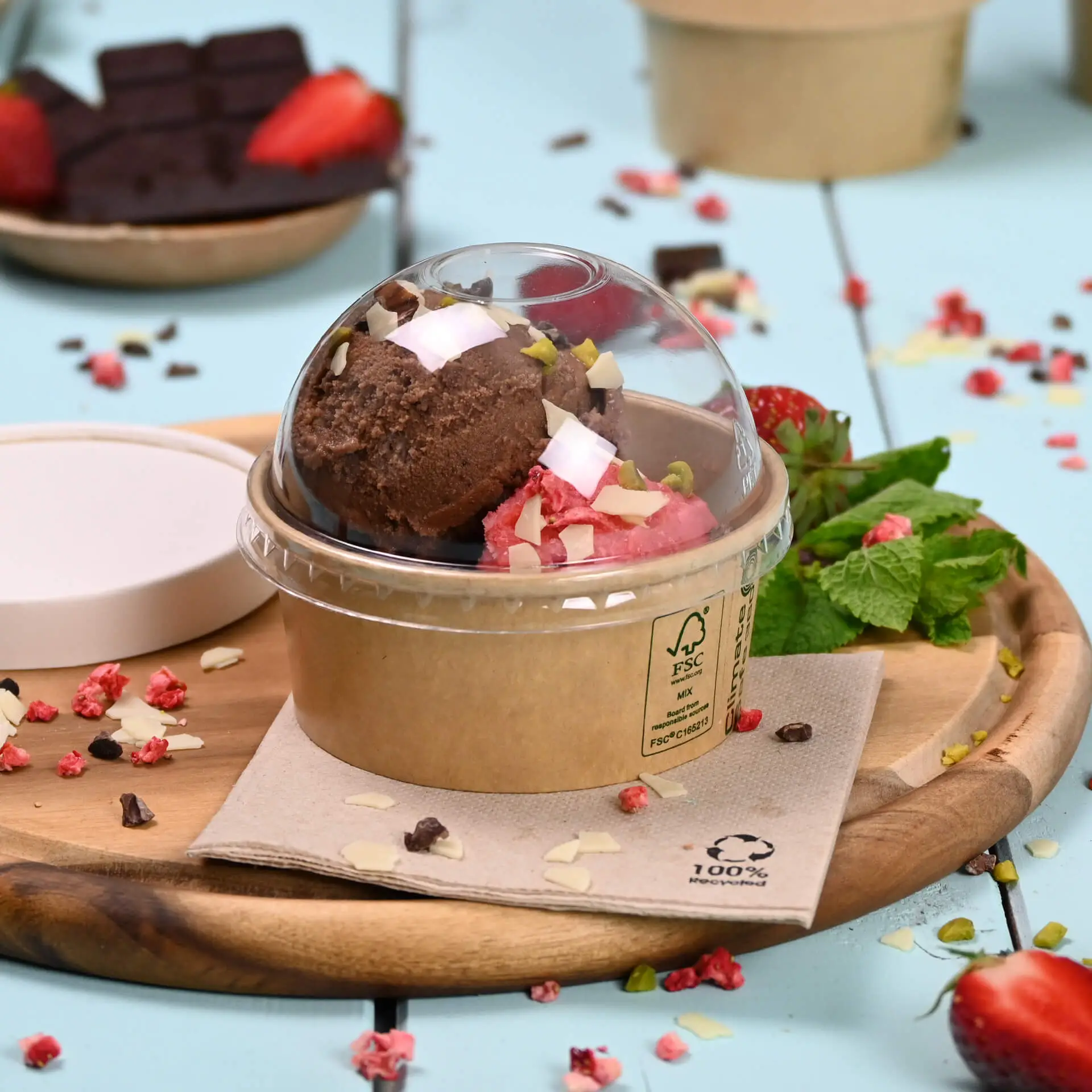 Okrągły kubek na wynos z dwiema gałkami lodów, jedną czekoladową i jedną truskawkową, posypany orzechami i wiórkami kokosowymi; przykrywka w kształcie kopuły na górze.