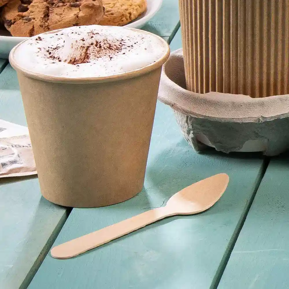 Brąski jednorazowy kubek z pianką i drewnianą łyżką na tealowym stole, z ciastkami w tle.