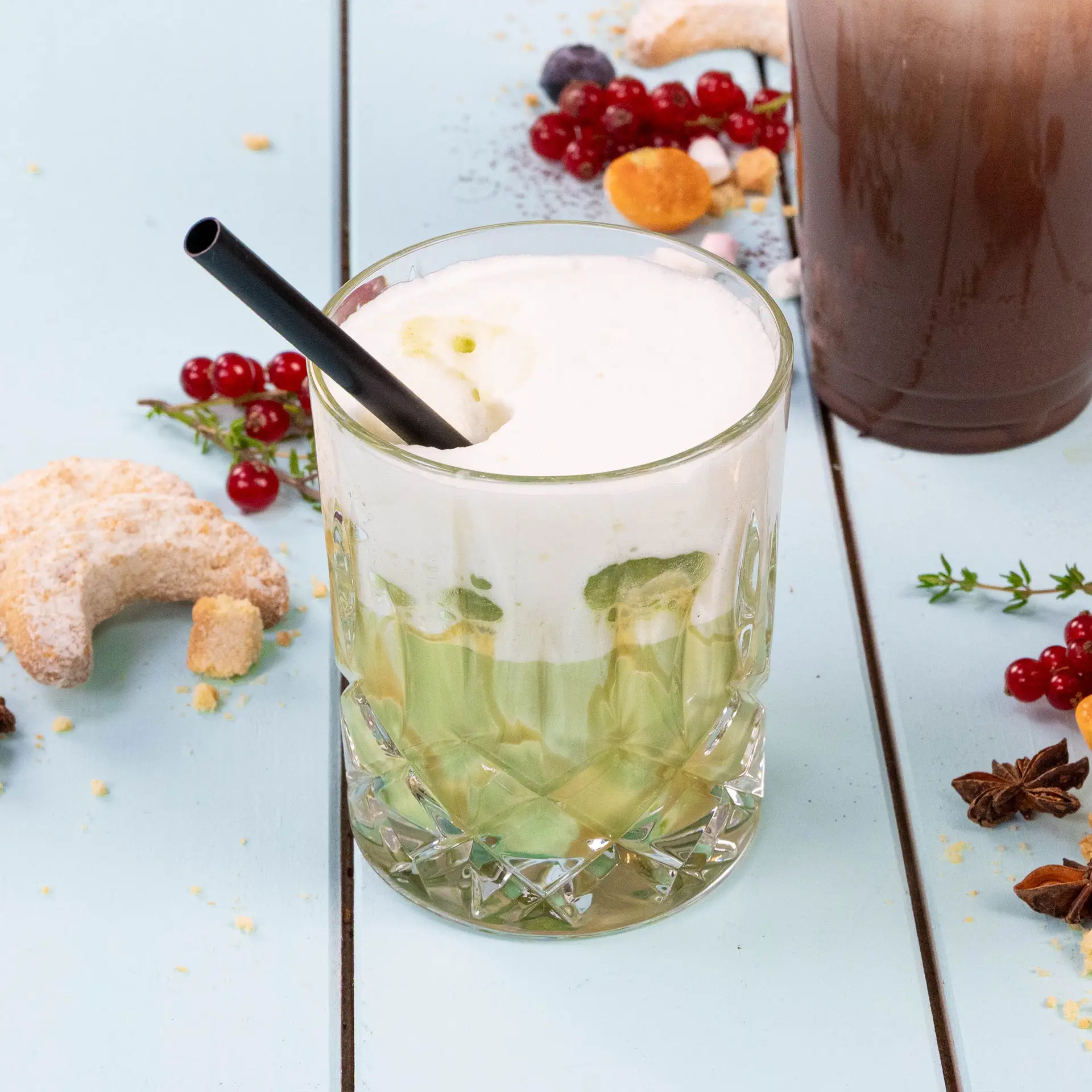 Szklanka wypełniona warstwowym zielonym napojem, zwieńczona puszystą białą pianką i czarną słomką; otoczona ciastkami i jagodami.