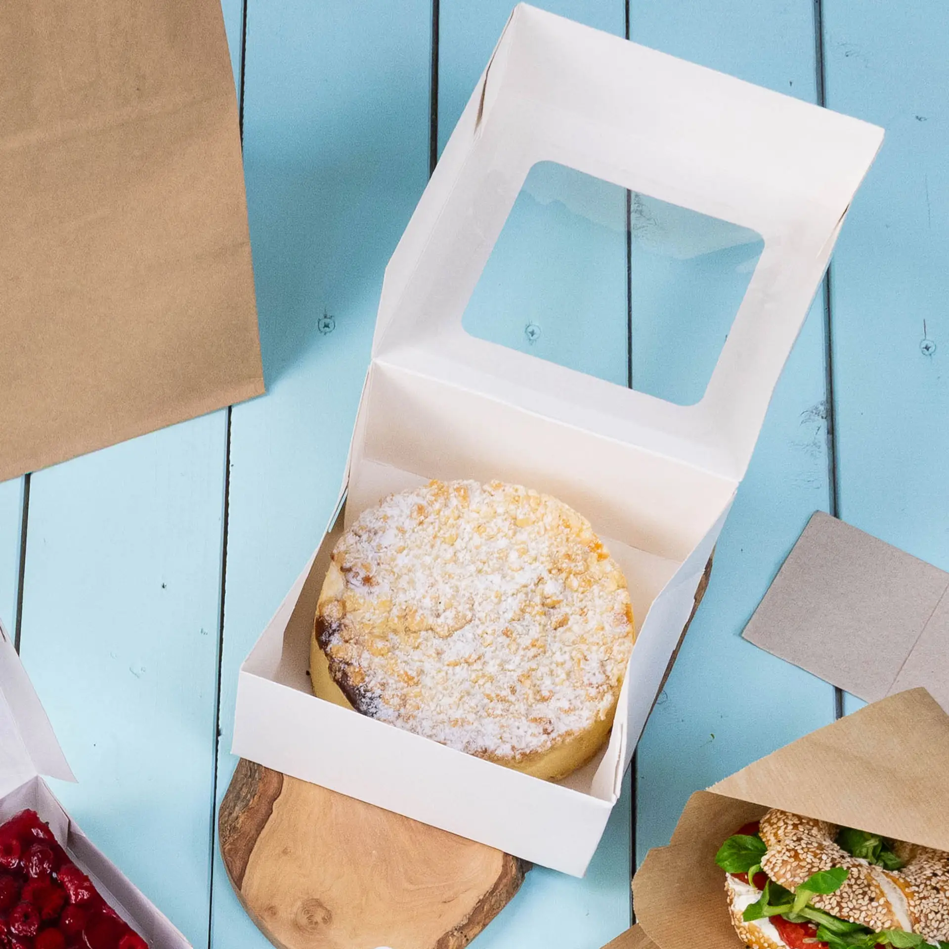 Pudełko na ciasto M w białym kolorze z otwartą pokrywką, w środku okrągłe ciasto, na drewnianej powierzchni z innymi artykułami spożywczymi w pobliżu.