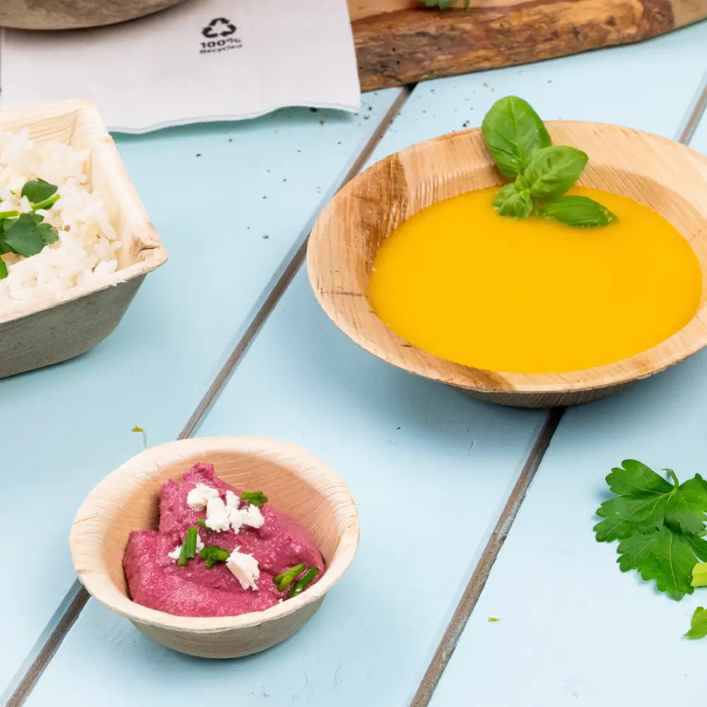 Bambusowa miska wypełniona żółtą zupą, ozdobiona liściem bazylii; mniejsza miska z różowym dipem posypana ziołami; ryż z ziołami w pobliskim pojemniku.