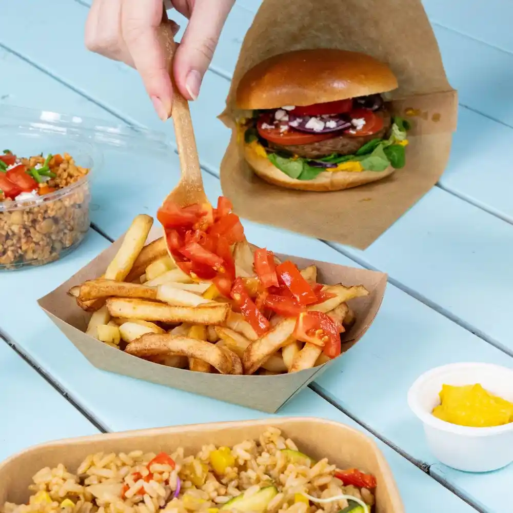 Ręka trzymająca drewnianą łyżkę dodaje pokrojone pomidory do porcji frytek na brązowej tacy; w tle jest hamburger i kubek ryżu.