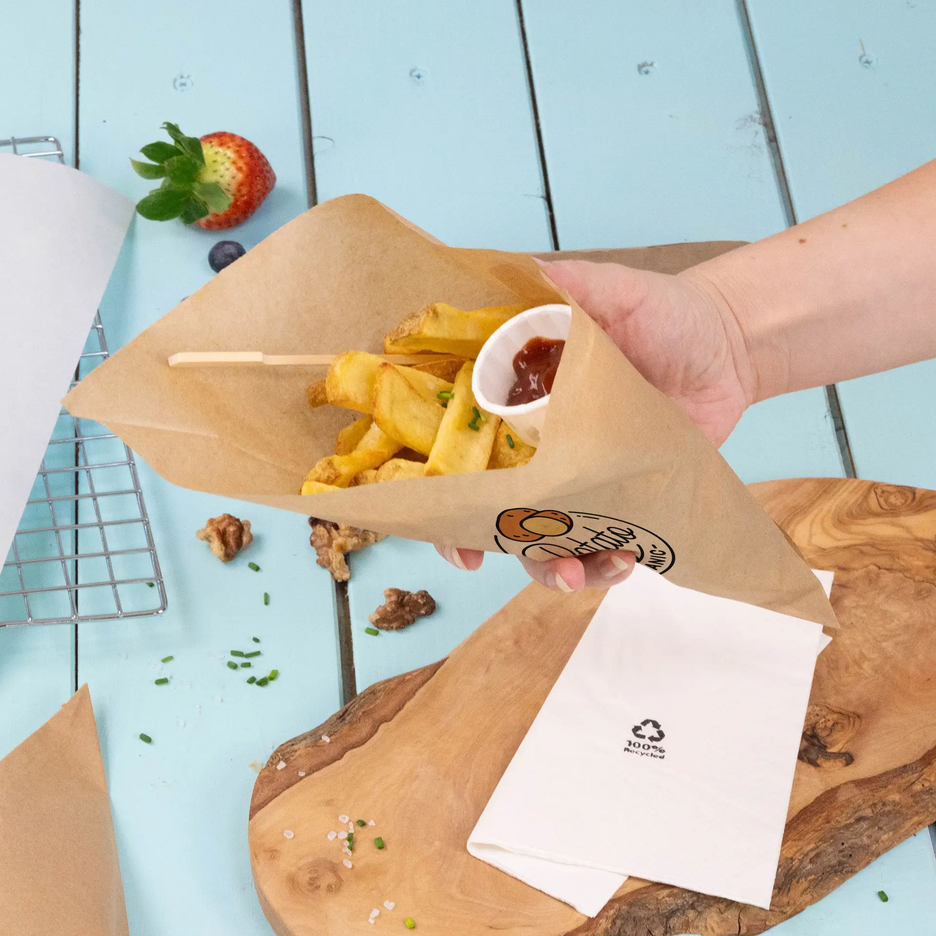 Ręka trzyma brązowy papierowy cone wypełniony frytkami i małą porcją ketchupu, na niebieskiej drewnianej powierzchni.