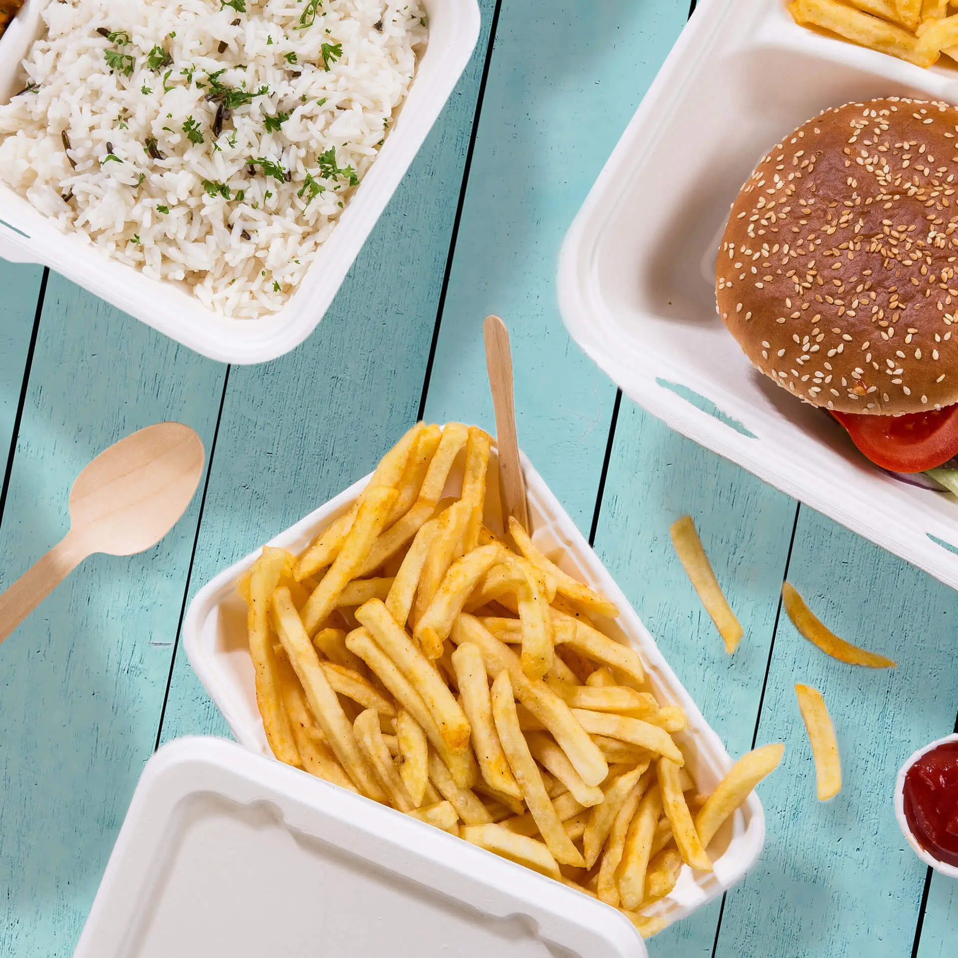 Biodegradowalna miska z trzciny cukrowej wypełniona frytkami, obok drewniana łyżka; ryż i hamburger w pojemnikach na wynos.